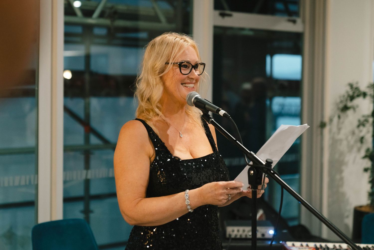 A speaker wearing a black sleeveless dress stands at a podium reading from papers during an indoor event.