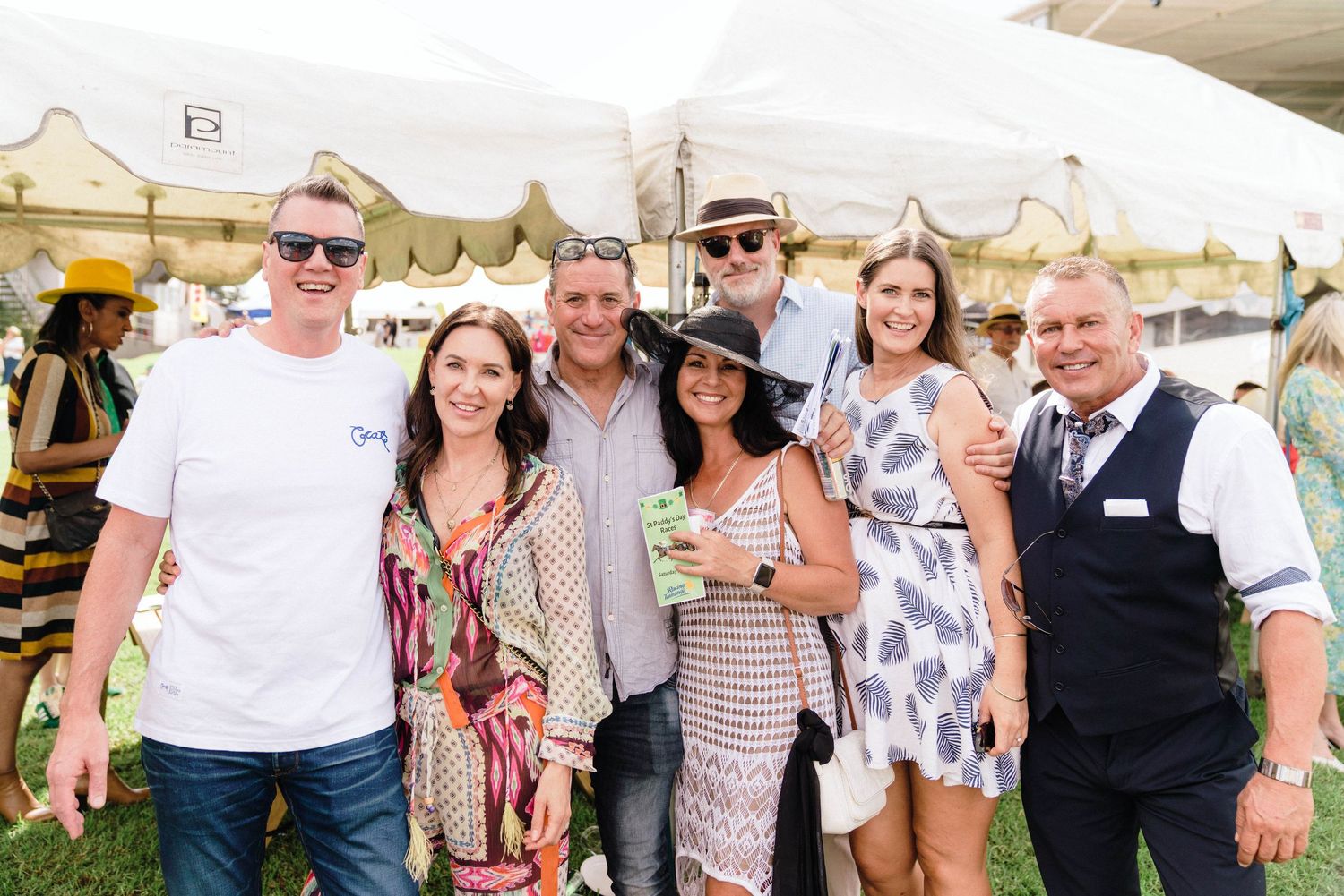 Group of friends in summer attire posing together at an outdoor event under white tents.