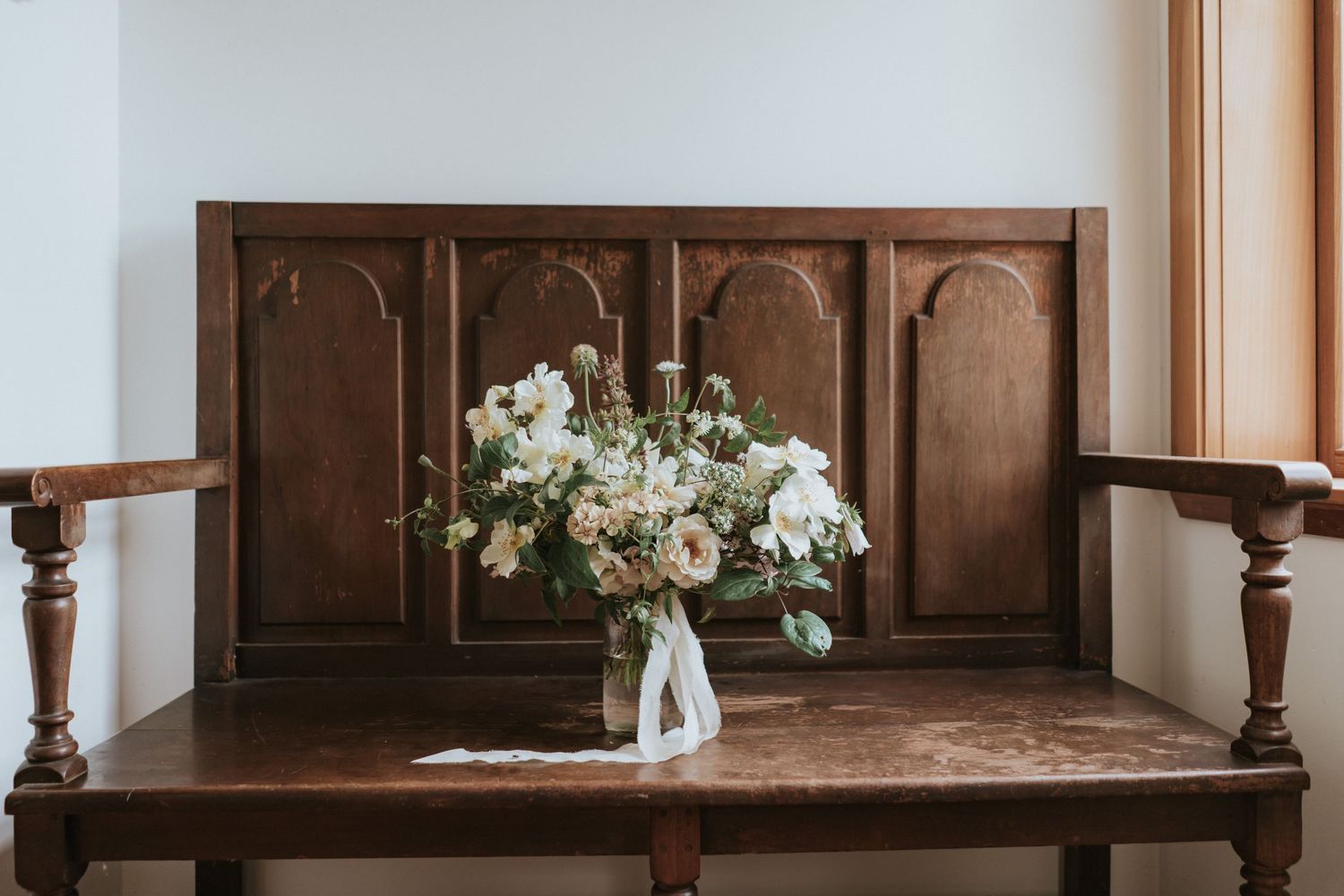 White floral arrangement in a clear vase sits on an antique wooden church pew with ornate carvings.