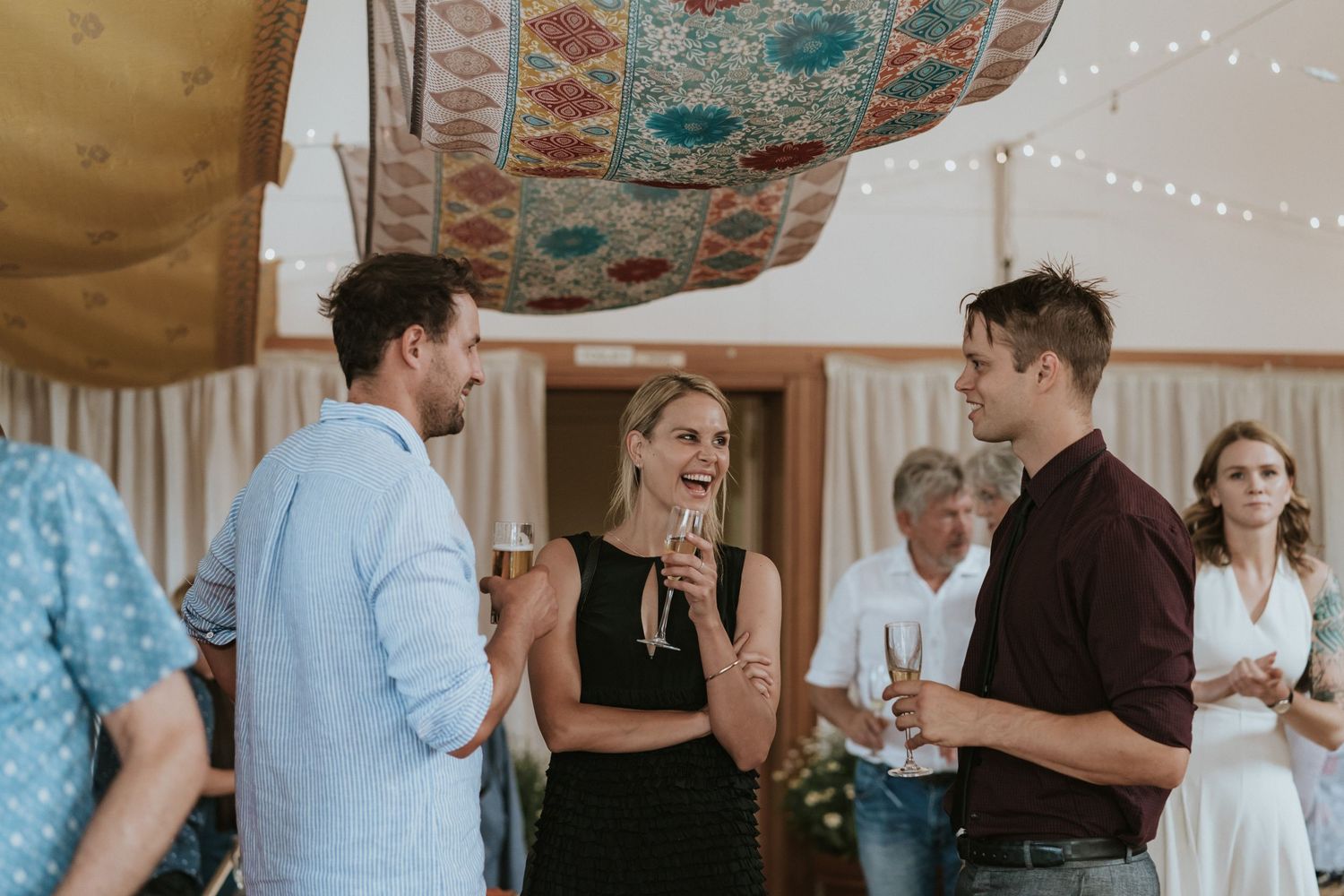 A group stands conversing at a casual indoor gathering with decorative tapestries hanging from the ceiling.