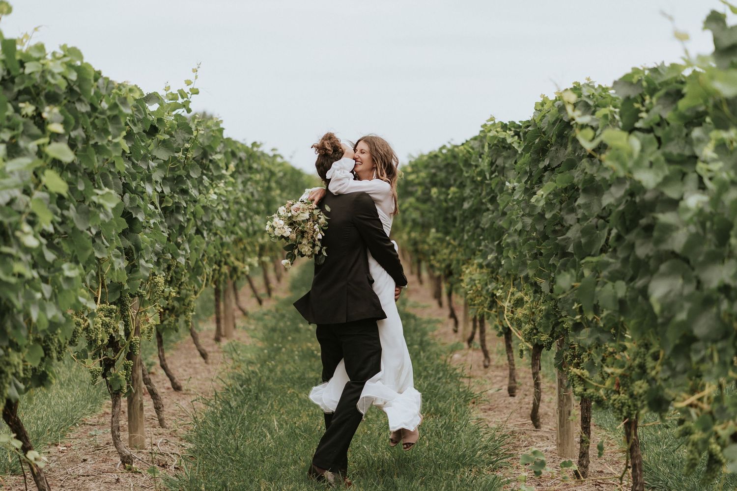 A romantic couple dances together in between rows of green grapevines at a scenic vineyard wedding.