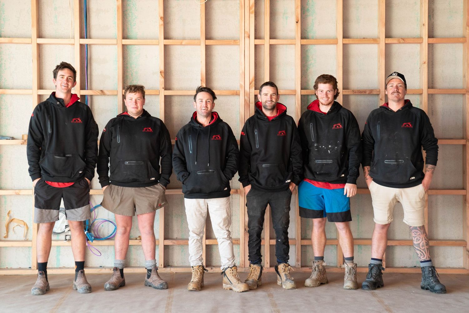 Six men in casual clothing standing together inside an unfinished construction space with wooden framed walls.
