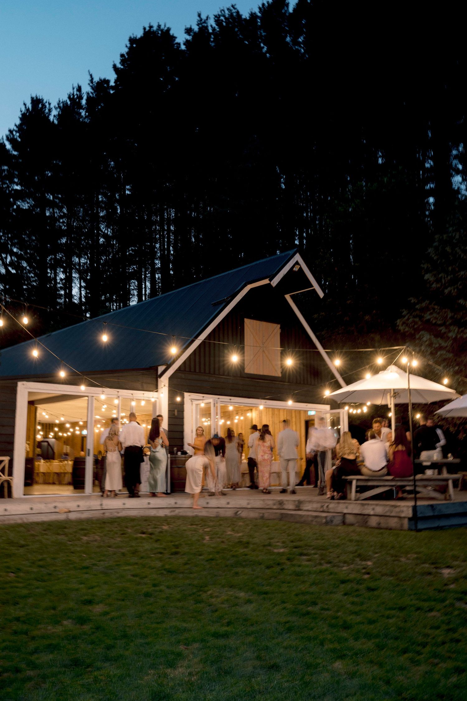 A modern barn-style wedding venue glows with string lights at dusk as guests gather outside.