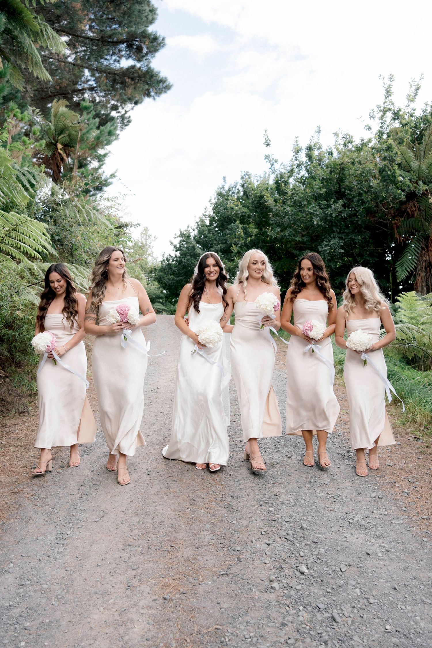 A group of bridesmaids in elegant cream-colored slip dresses walk together on a dirt path surrounded by greenery.