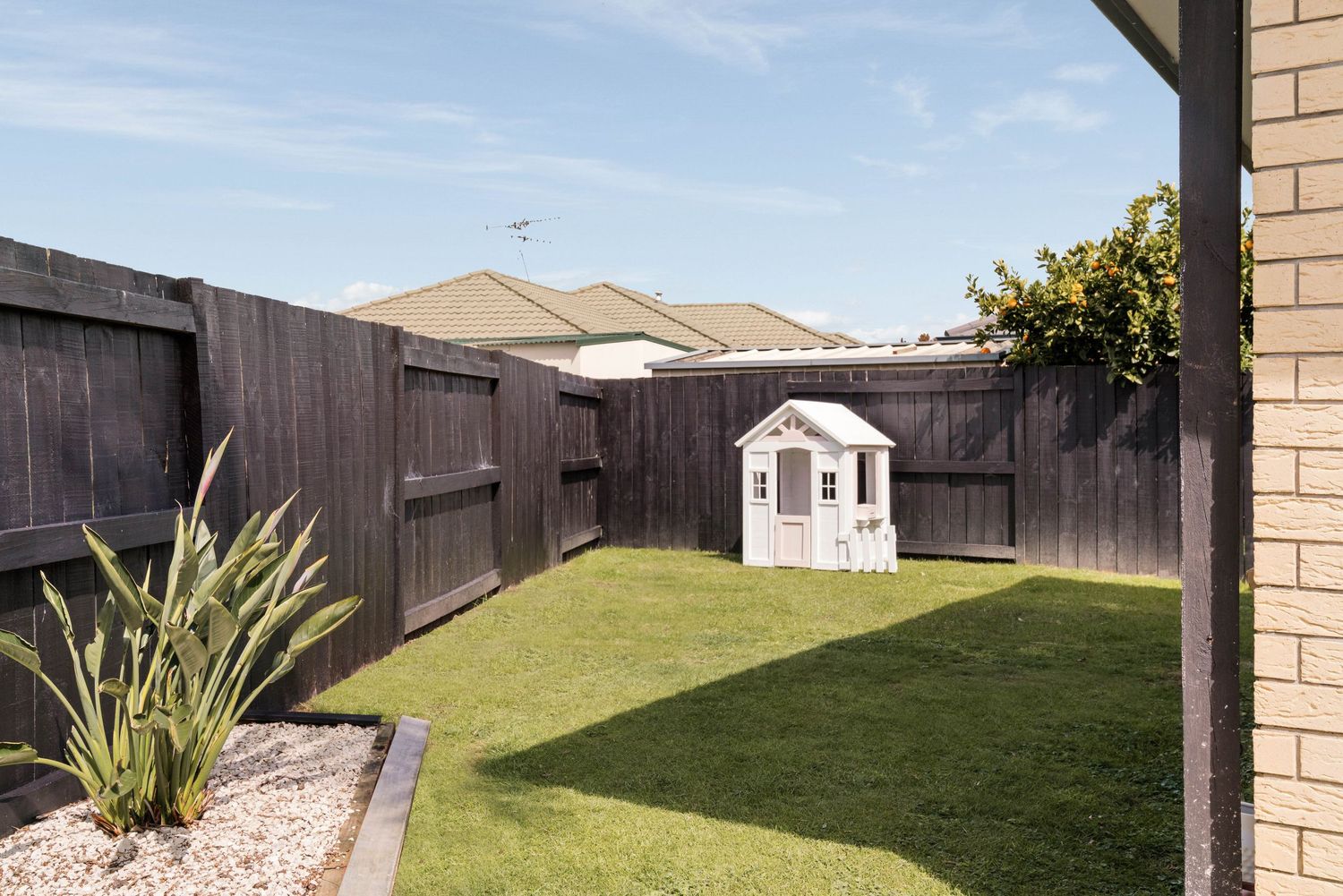 Small playhouse in landscaped backyard with artificial turf and wooden privacy fence.