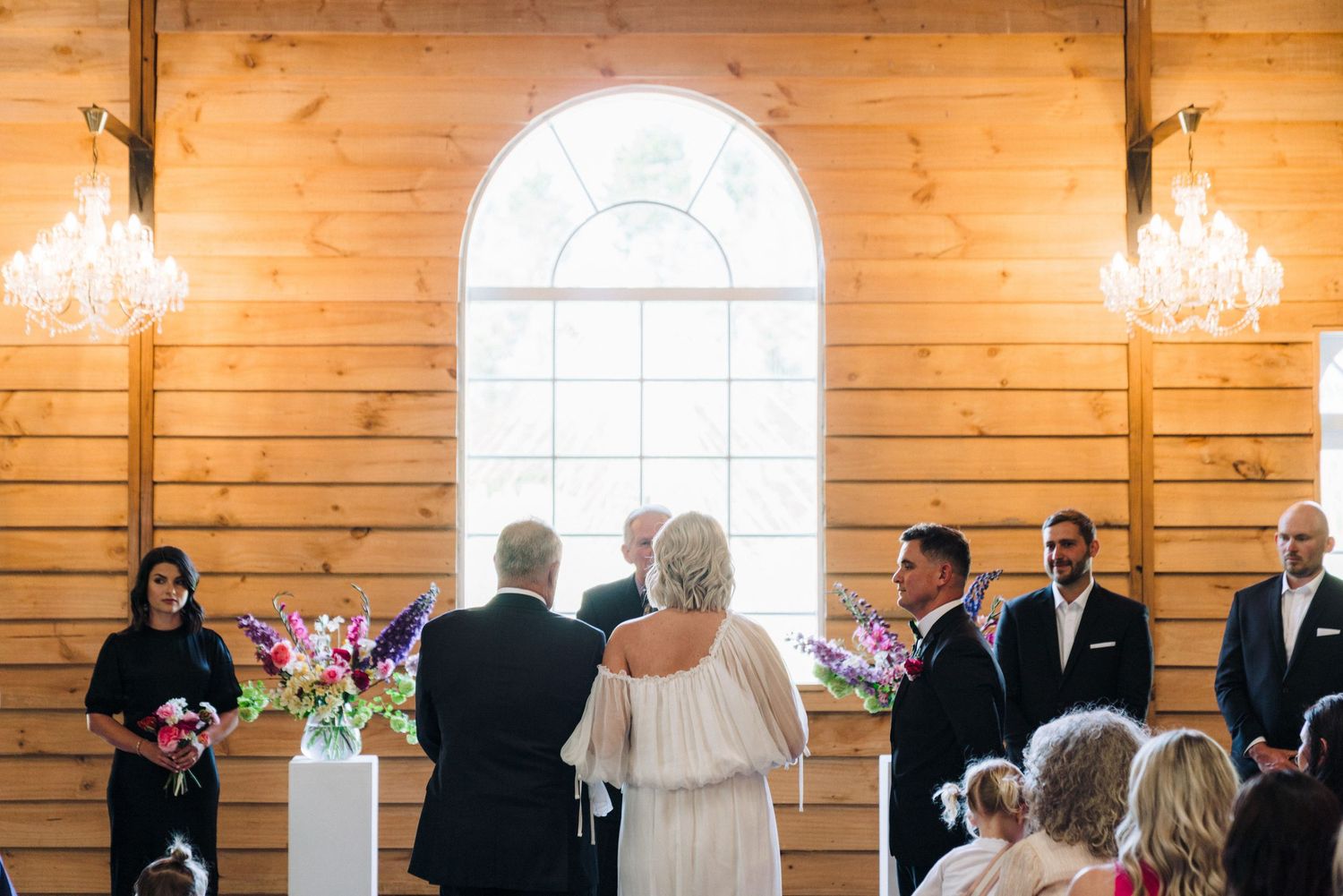 Warm wooden barn venue with chandeliers and guests gathered for a wedding ceremony.