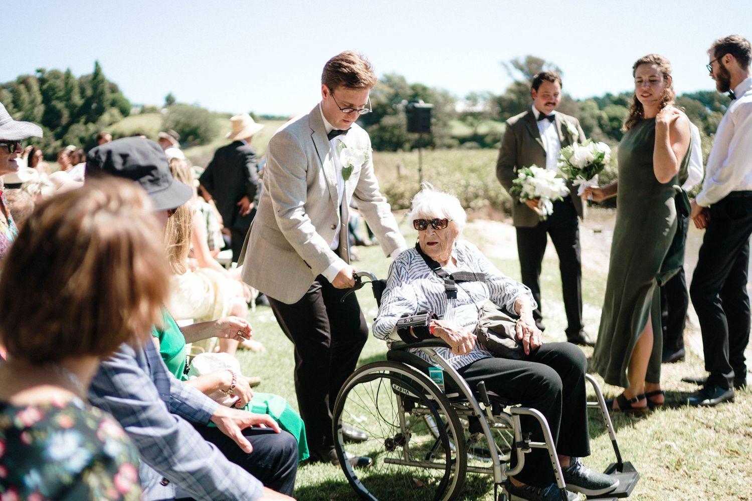 Wedding guests gathered outdoors around a wheelchair at a sunny outdoor celebration.