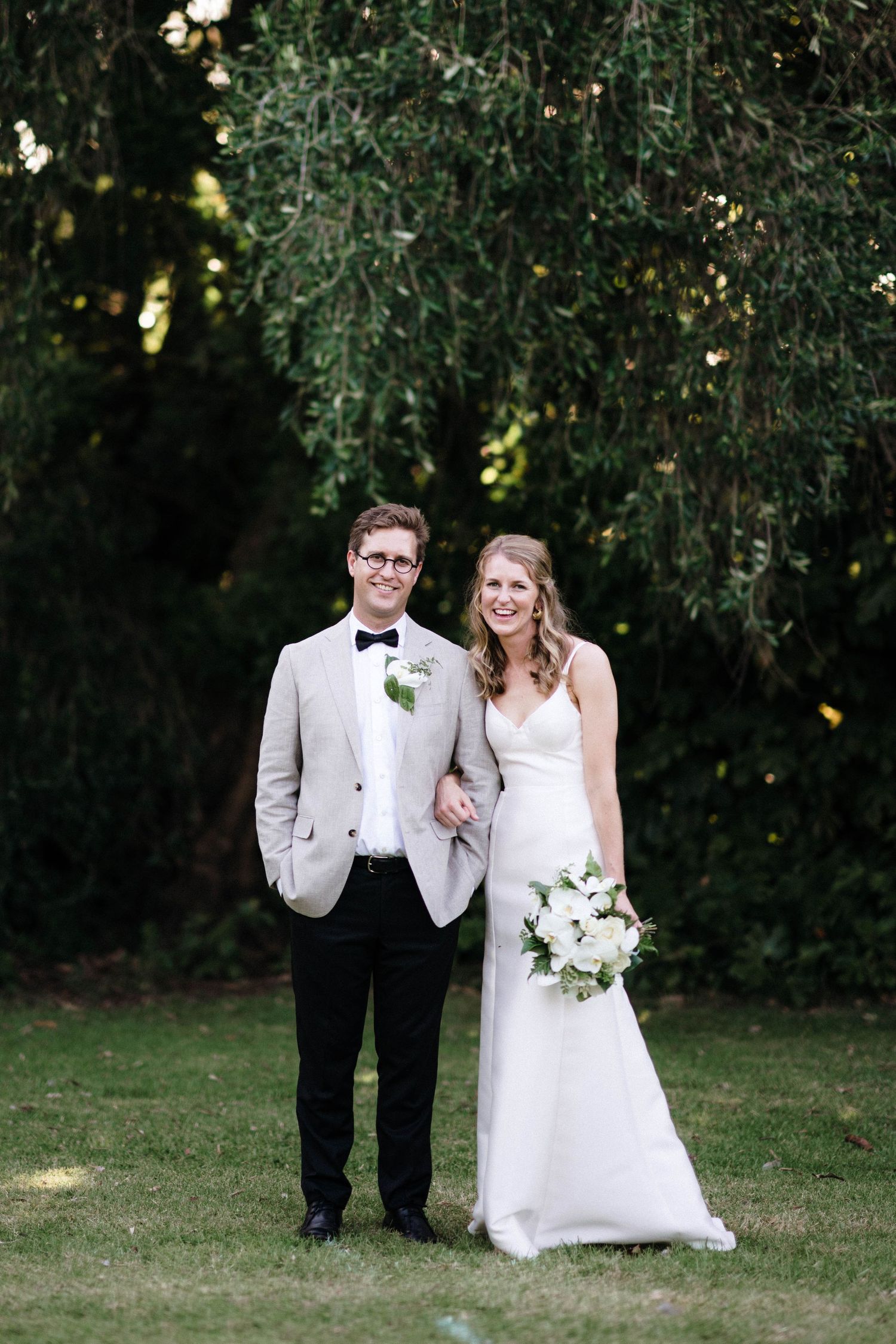 Elegant couple in formal wedding attire pose together on grass under trees.