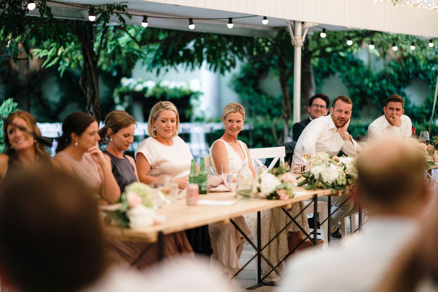 Reception guests seated at a long table decorated with floral arrangements and candles under an outdoor tent.