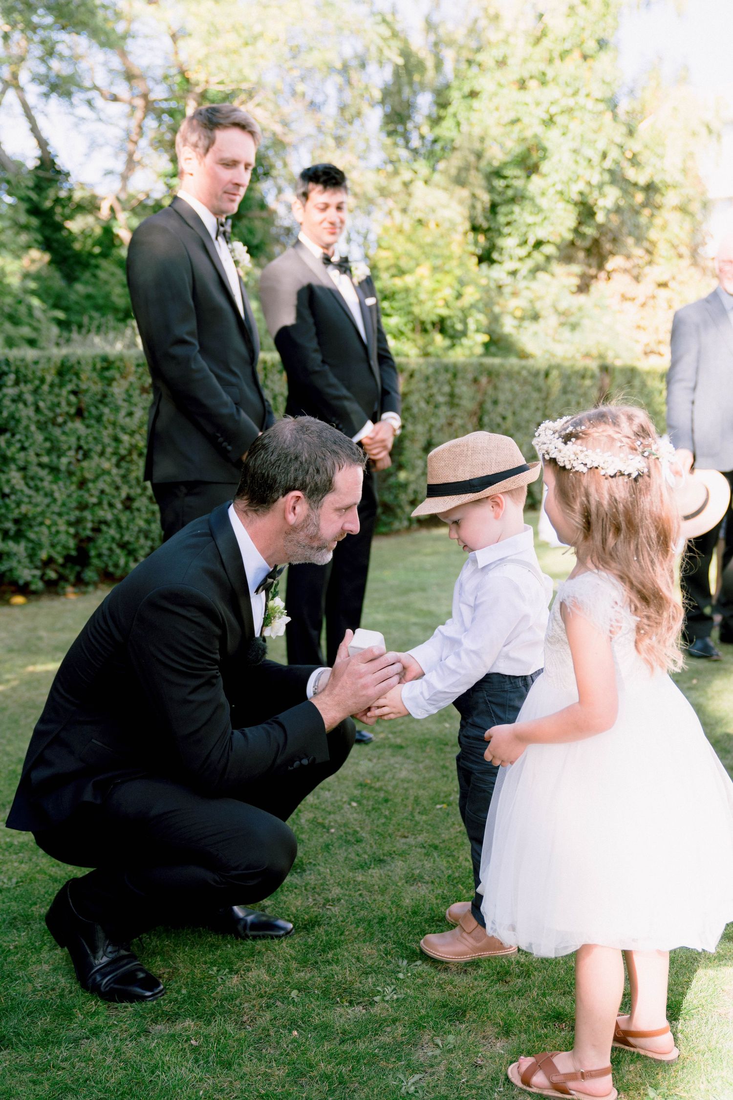 A tender moment at an outdoor wedding as a man in a black suit crouches down to greet young children on a lawn.