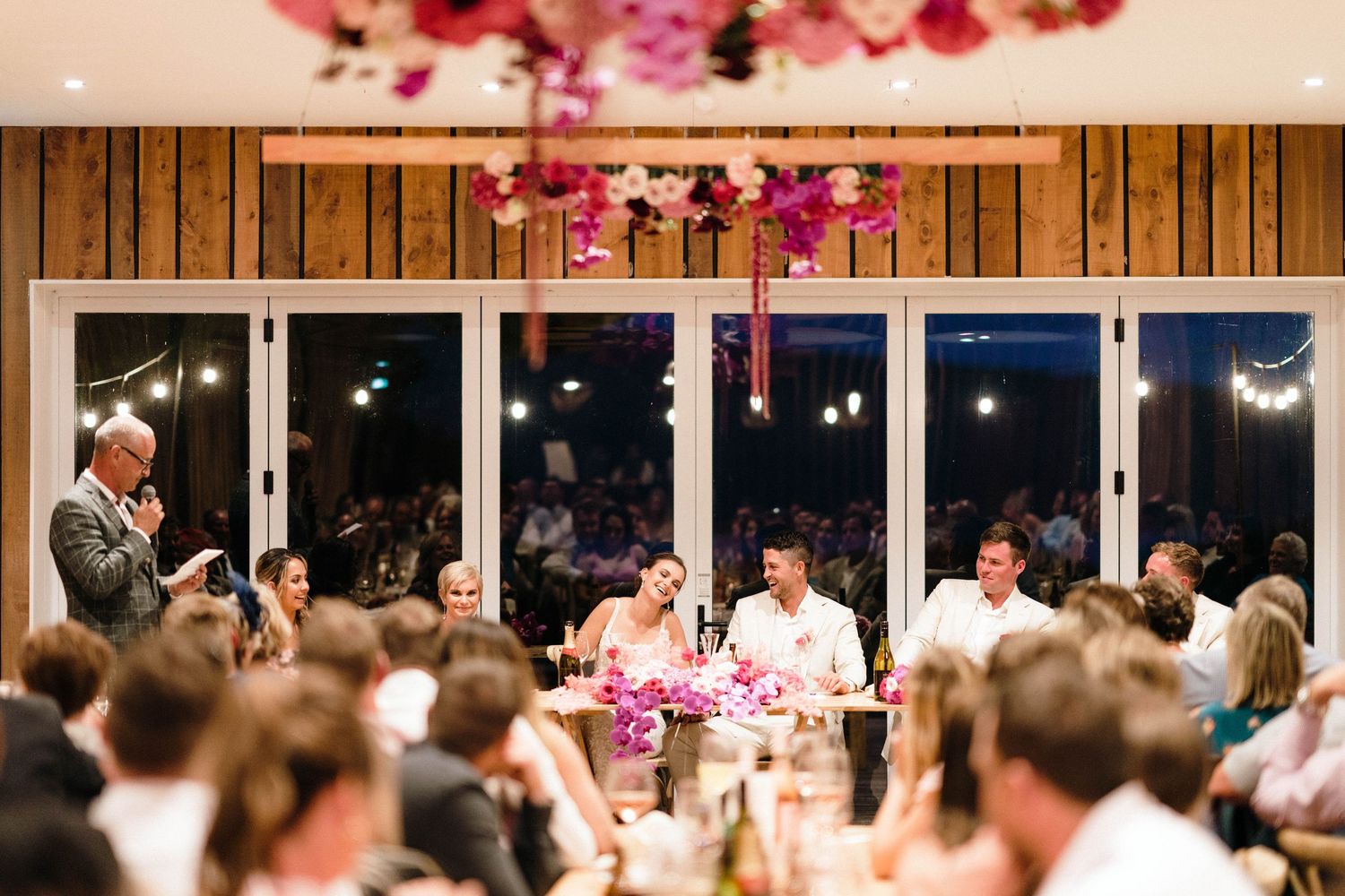 Reception venue with wooden walls and pink floral ceiling decorations during evening celebrations.