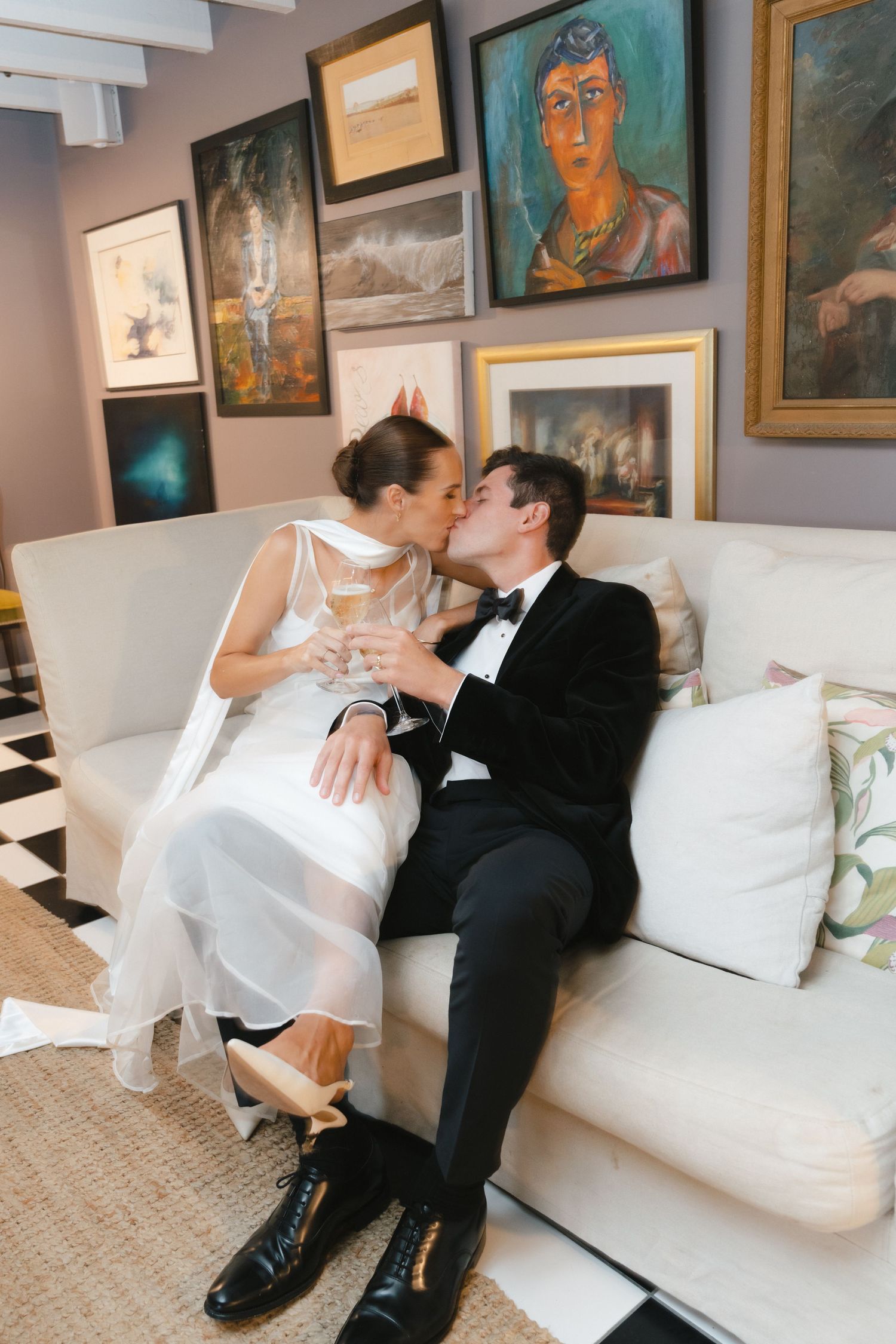 A couple in formal wedding attire share an intimate moment on a white couch with framed artwork displayed on the wall behind them.