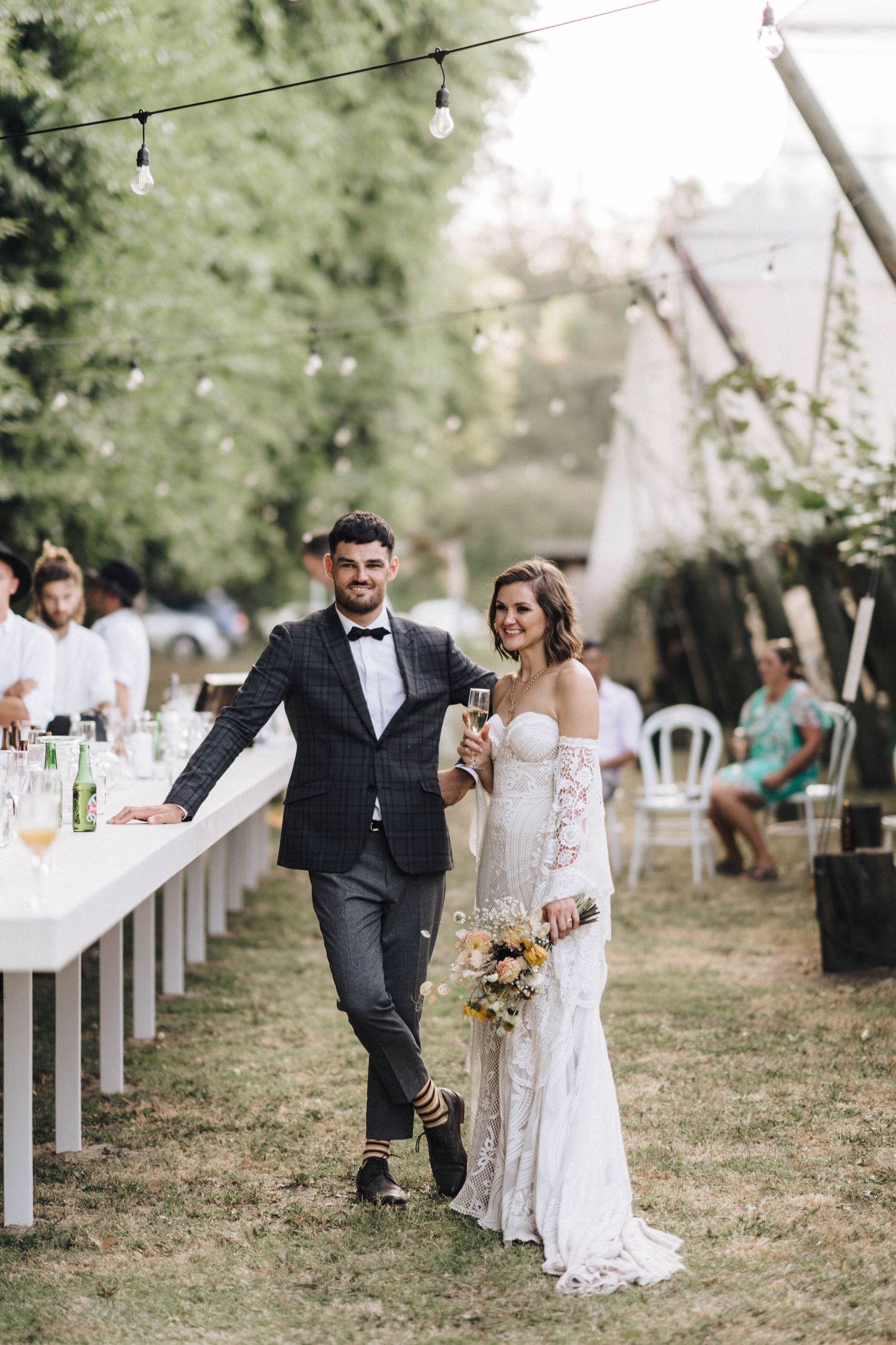 Newlyweds walk together at outdoor wedding reception with long banquet tables and string lights in background.