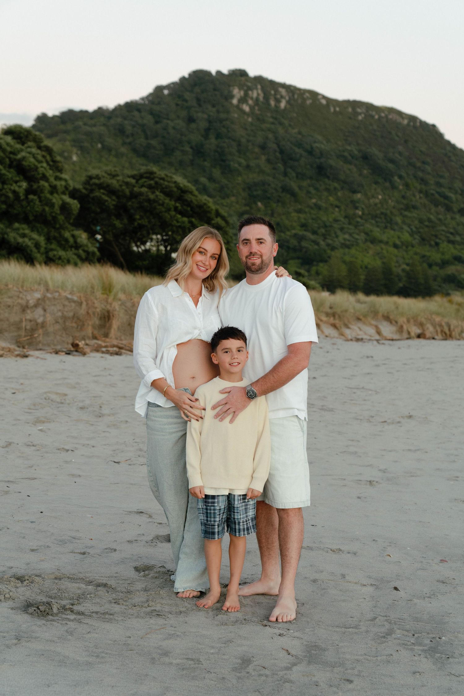 Pregnant woman in white shirt poses with partner and young son on sandy beach with green hills backdrop. Photographer Bryony at Newfound.