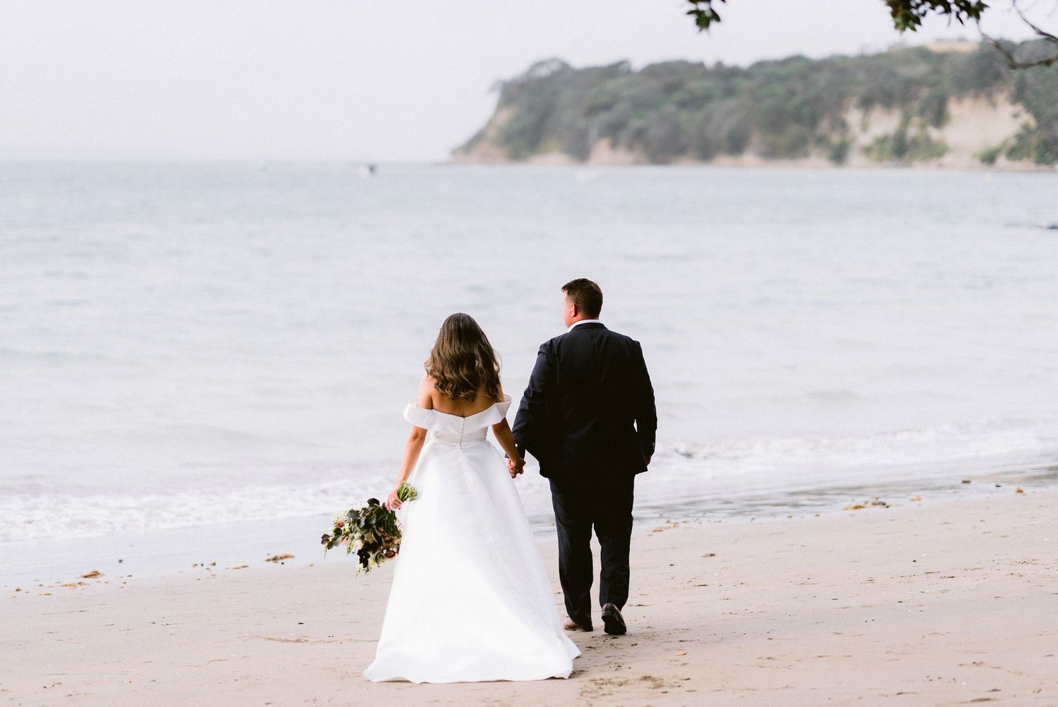 Beach wedding couple walking along scenic coastline with cliffs in background.