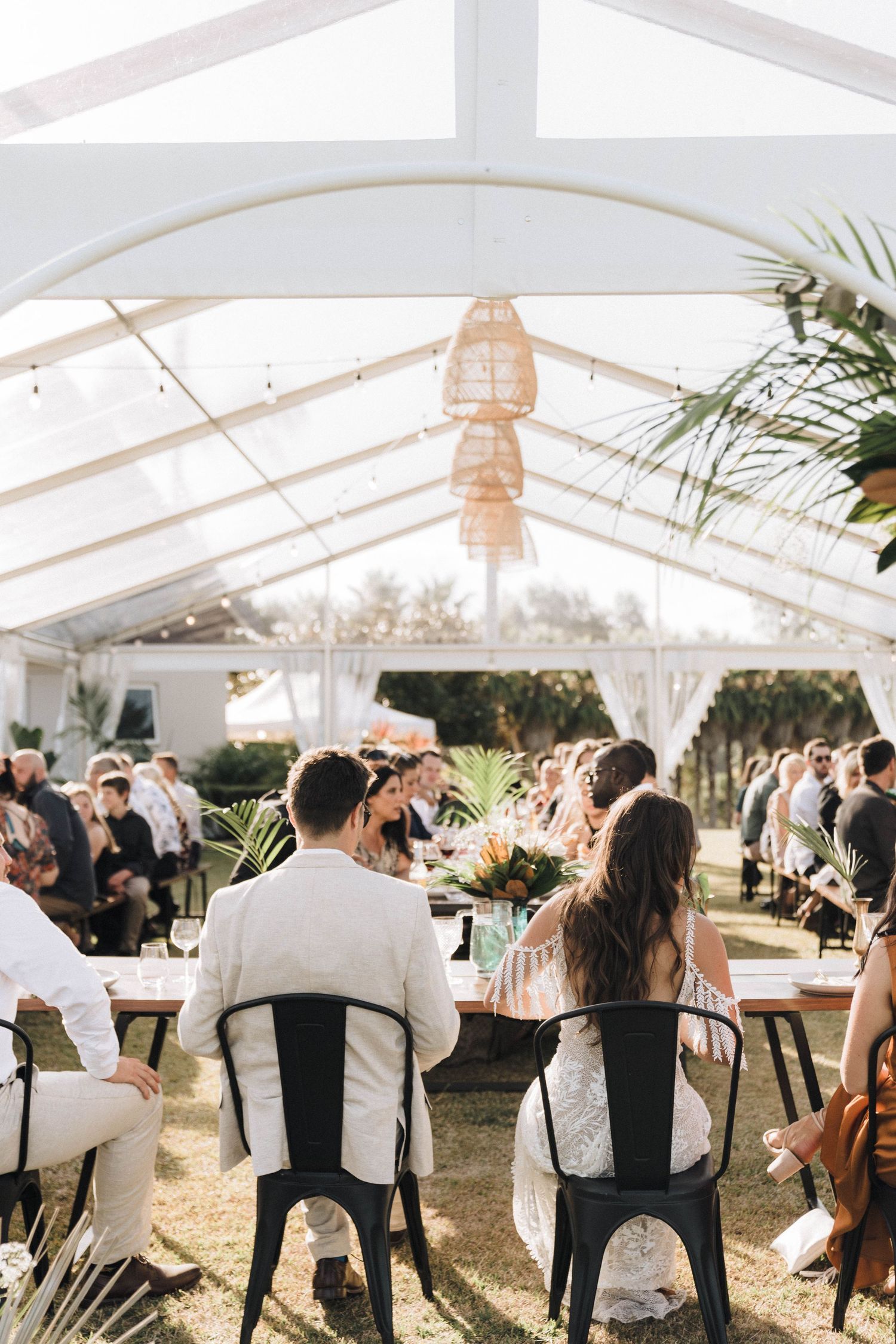 Wedding reception setup under clear tent with hanging lights and tropical decor.