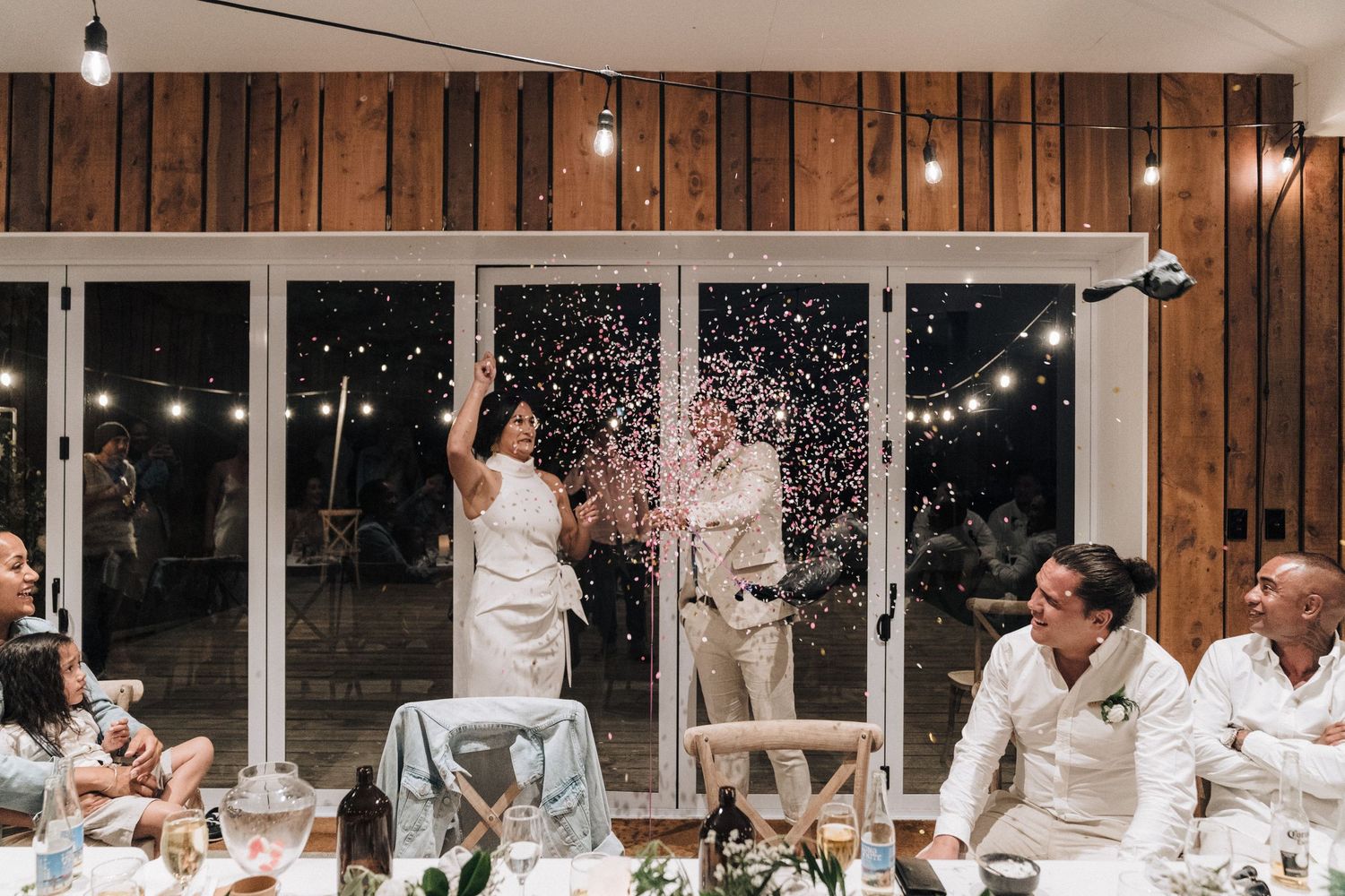 Wedding reception moment with champagne spray inside modern venue with string lights and wooden walls.