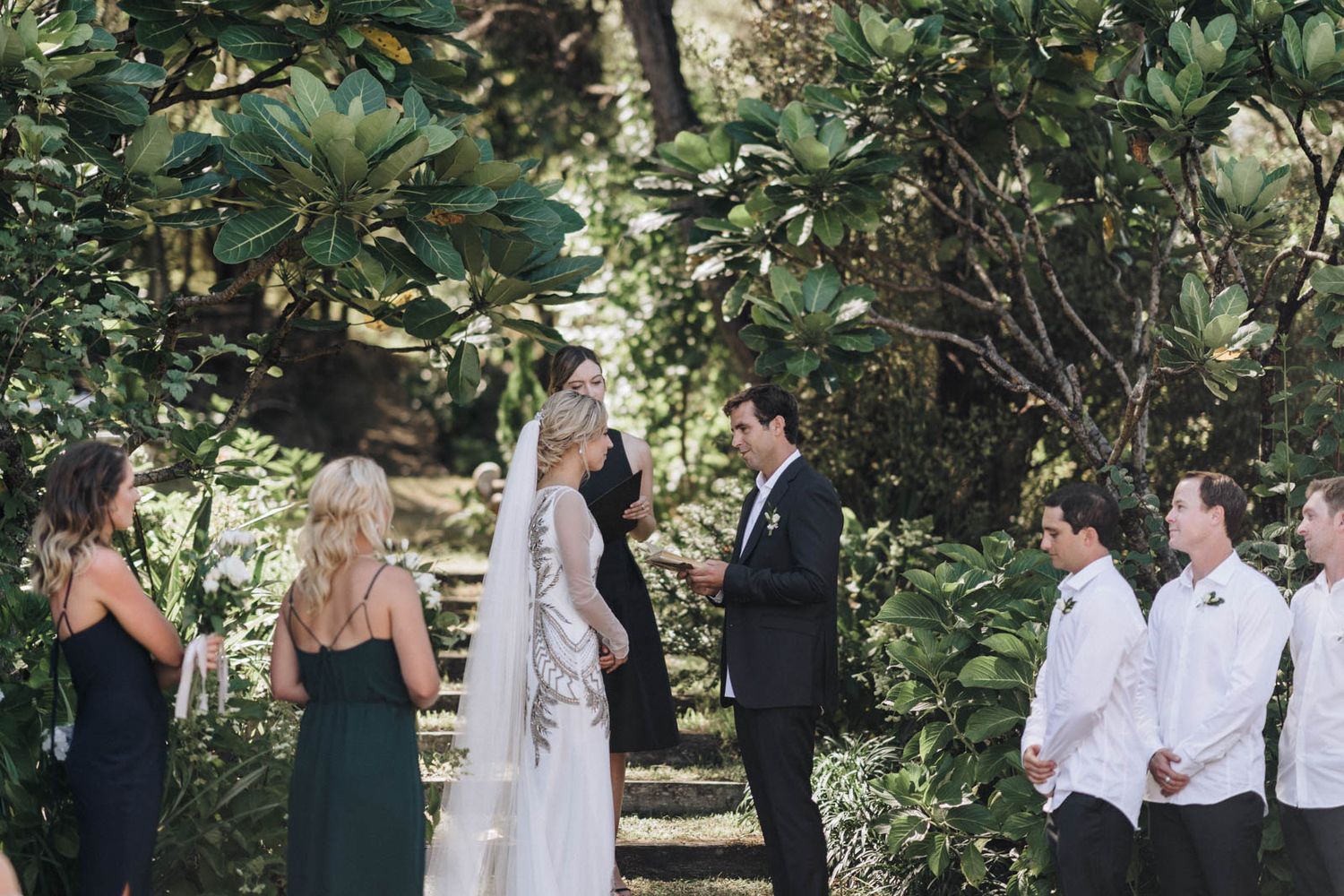 An outdoor wedding ceremony takes place in a lush garden setting surrounded by tropical foliage and greenery.