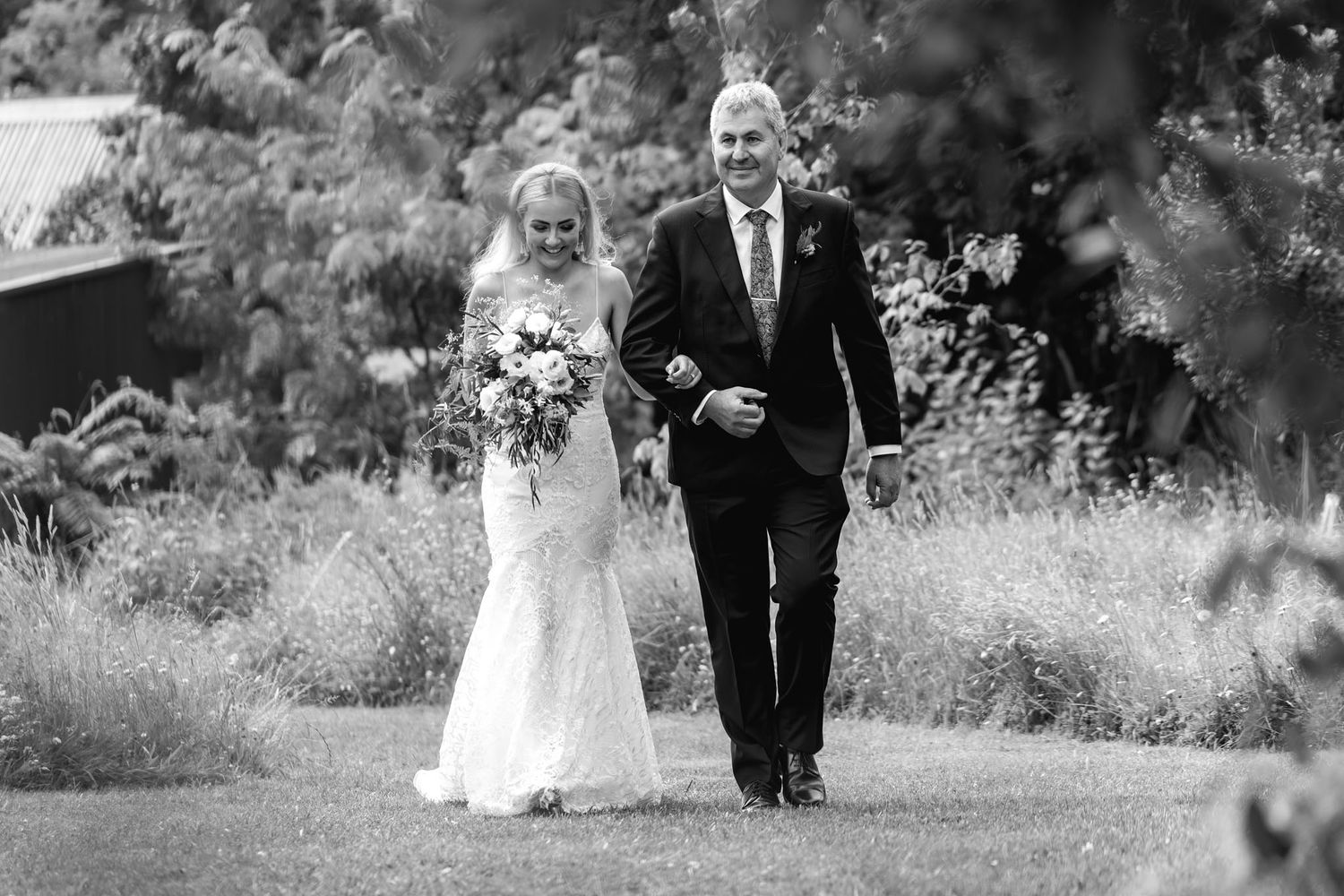 A serene outdoor wedding photoshoot shows the couple walking together through a lush garden setting in black and white.