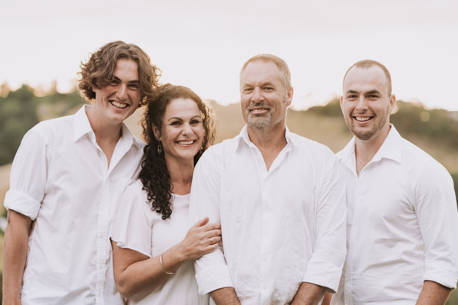 A cheerful family wearing white clothing poses outdoors at sunset during a casual photo session.