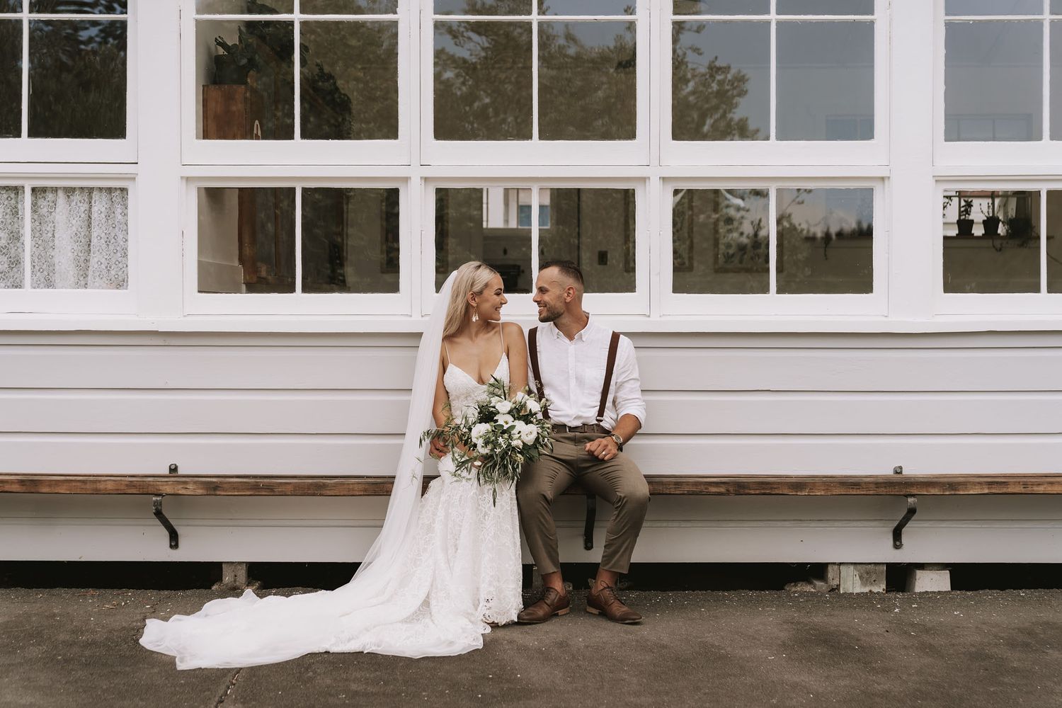 A rustic wedding scene with a couple in wedding attire sitting together on a white building's steps holding a floral bouquet.
