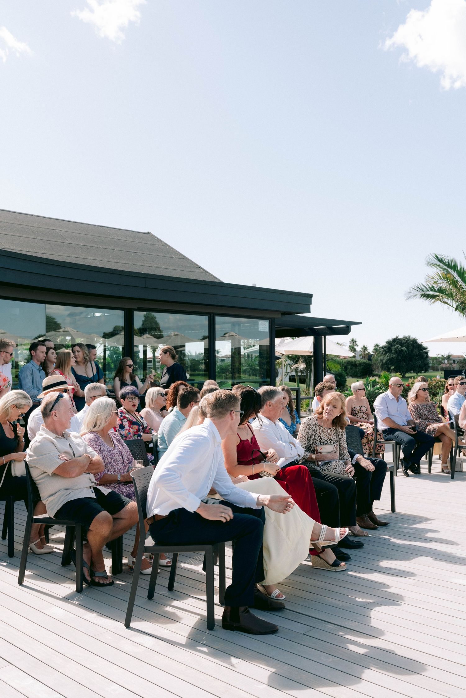 Wedding guests gather under covered outdoor patio seating area during daytime celebration.