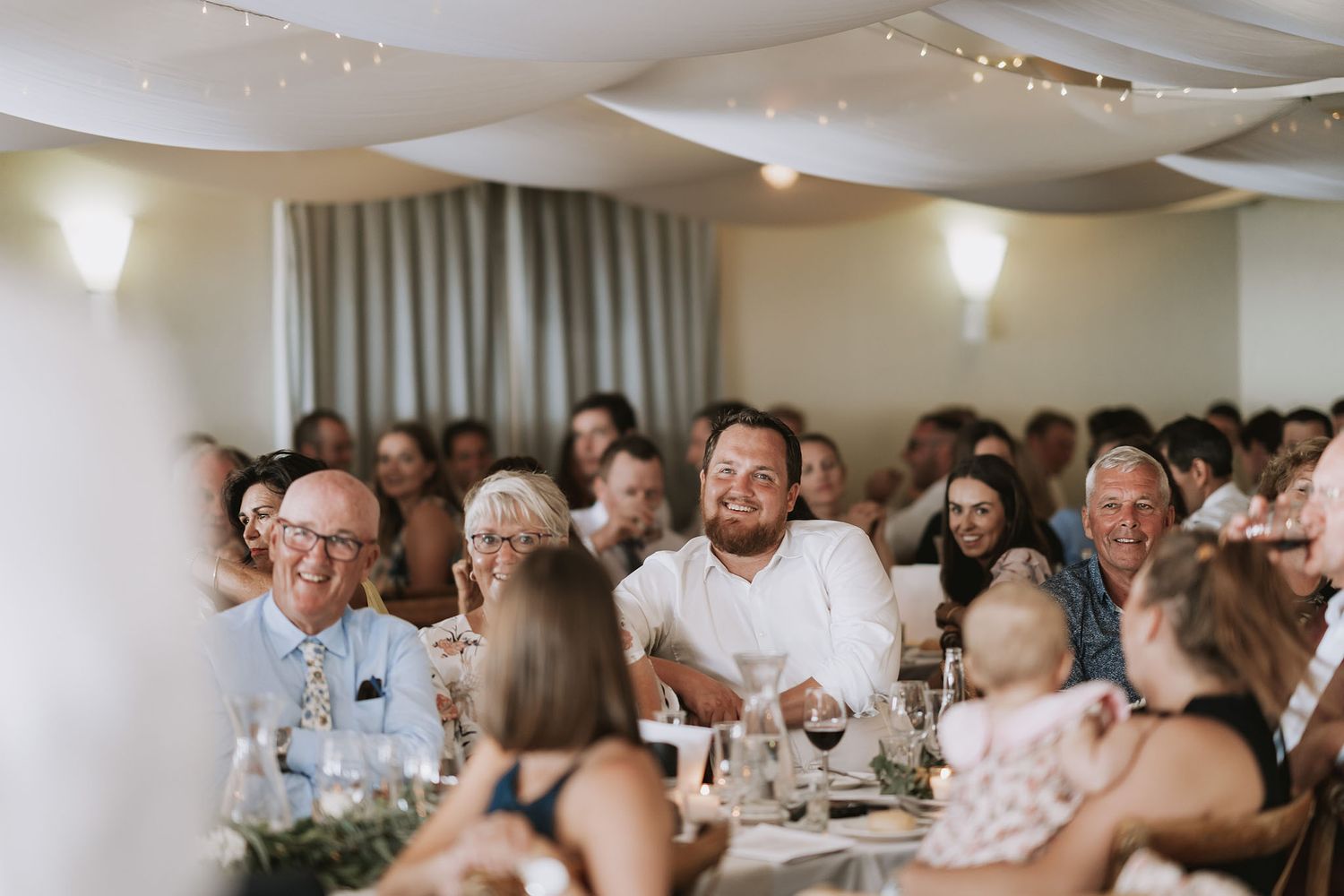 Guests enjoying drinks and conversation at a lively indoor reception with white tablecloths and warm lighting.