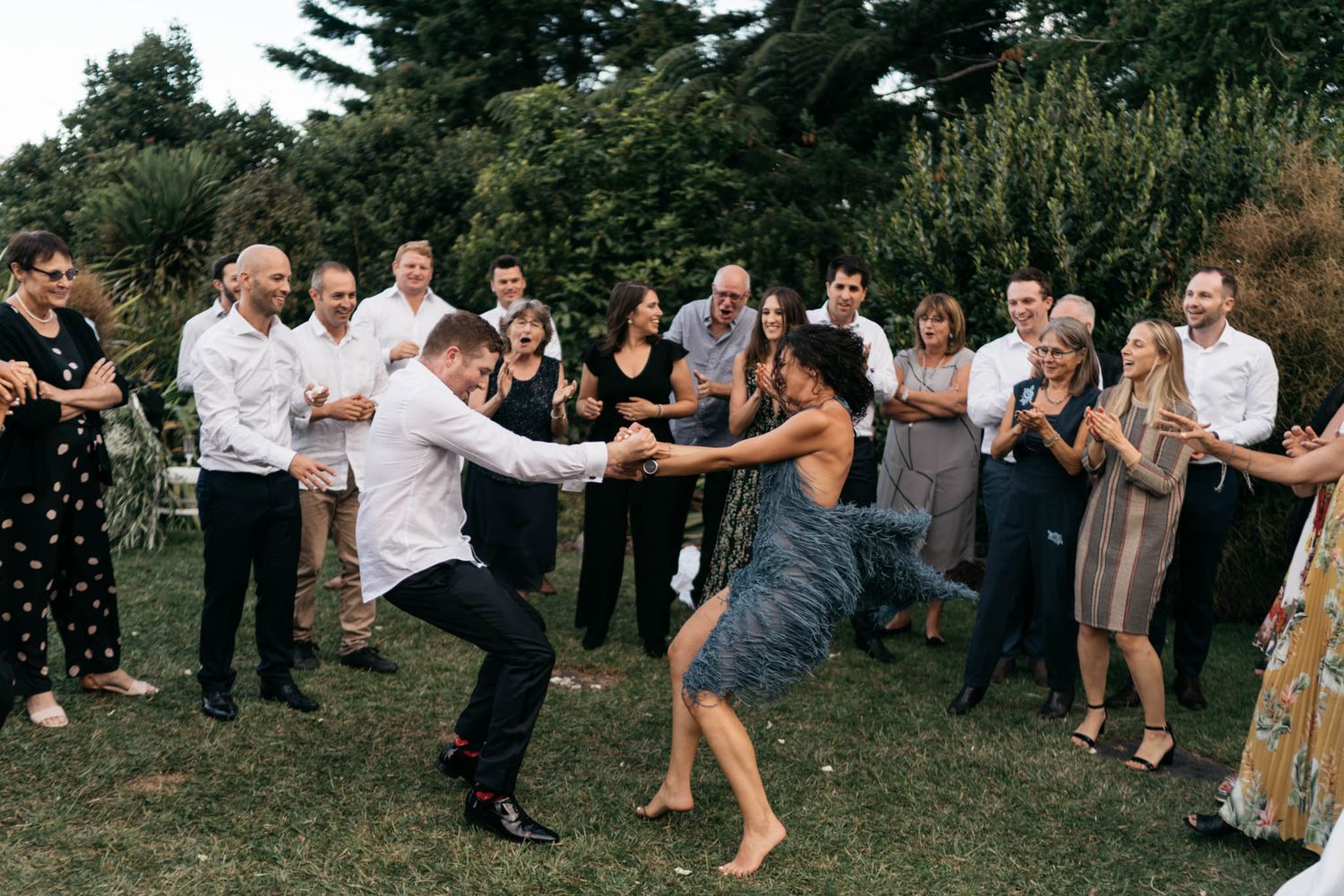 Wedding guests dance and celebrate on a grassy lawn during an outdoor reception while others watch and cheer.