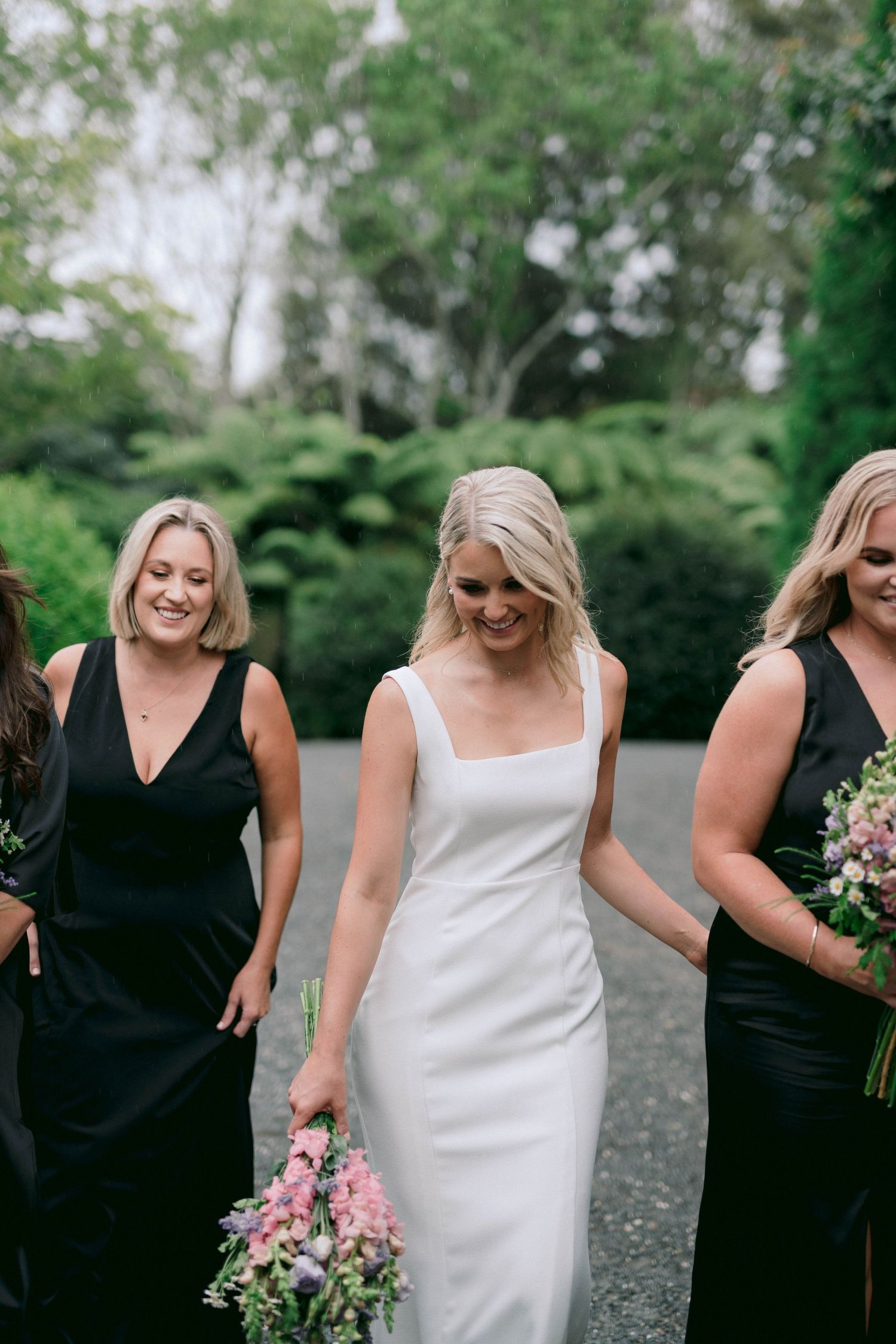 Bride in sleek white dress walking with bridesmaids in black dresses holding pink floral bouquets.