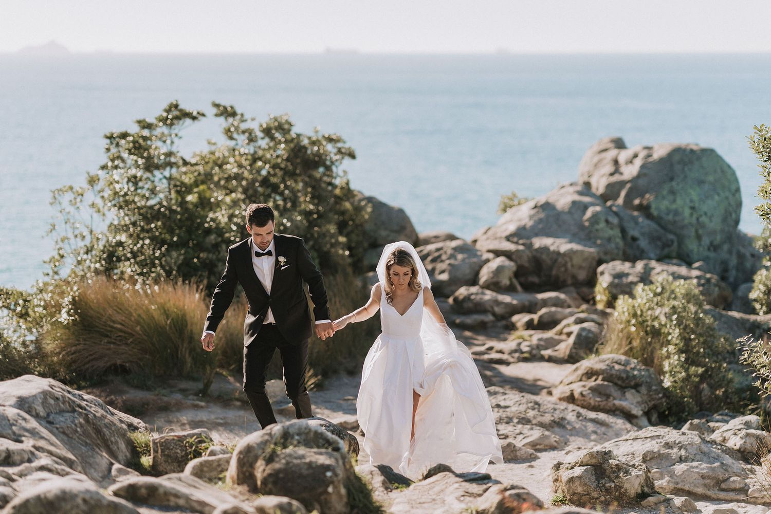 Newlywed couple walking together on coastal rocky cliffs with ocean view on sunny day.