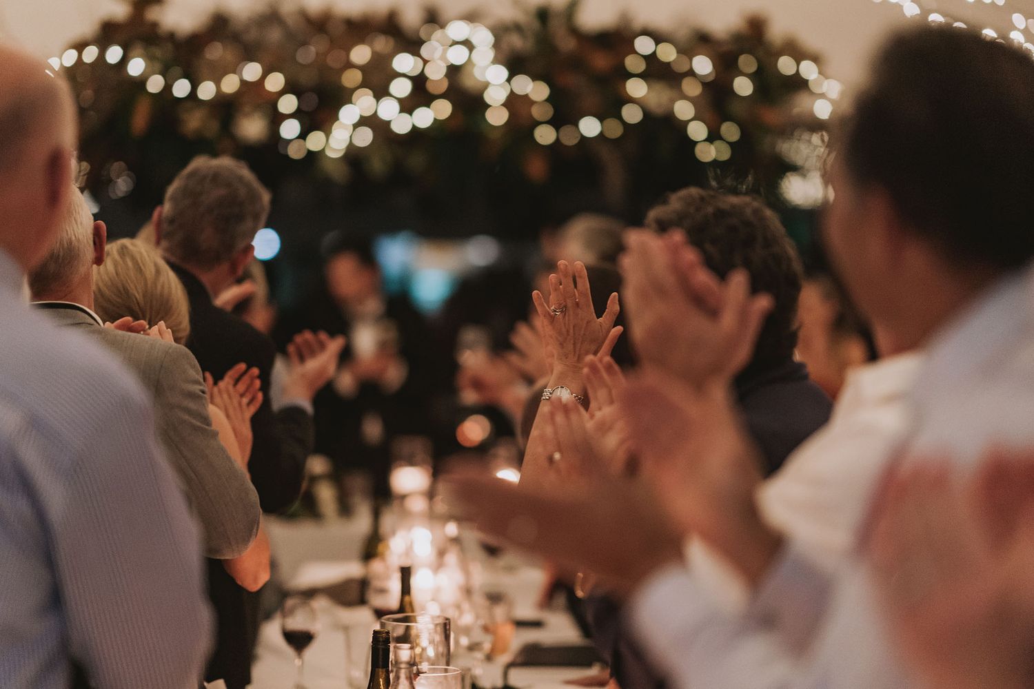 Wedding guests clap and celebrate at a candlelit reception venue decorated with twinkling lights overhead.