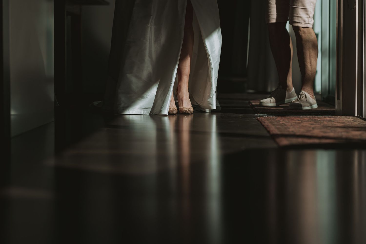 Bare feet and legs stand in a dimly lit hallway with shadows and reflections on a hardwood floor.
