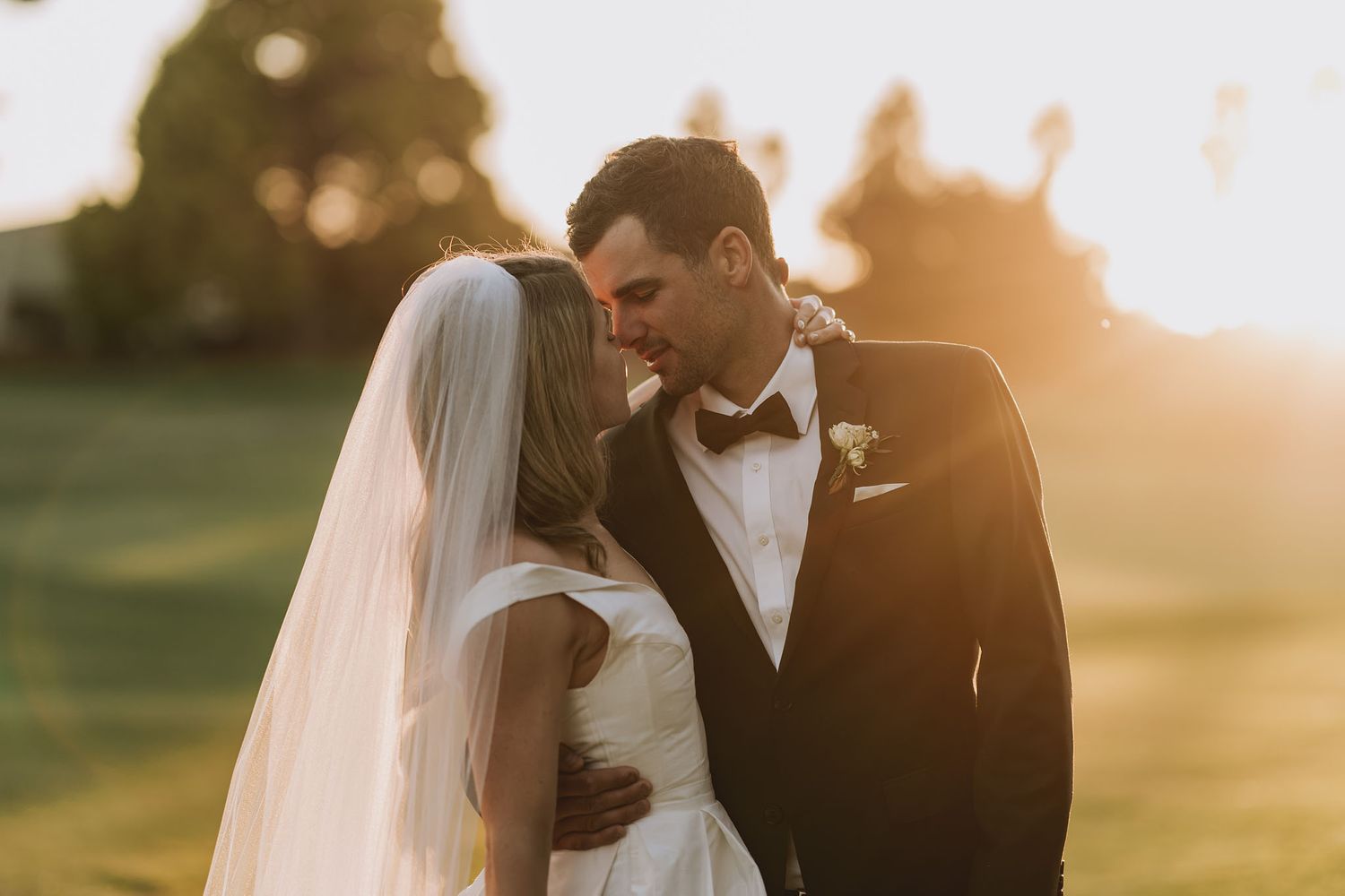 Wedding couple embracing at sunset in golden light on grassy field with dreamy bokeh background.