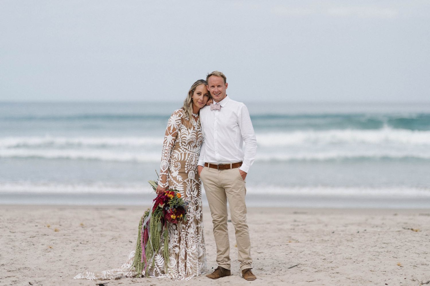 A coastal beach wedding ceremony with guests and decor set up on the sand next to ocean waves.