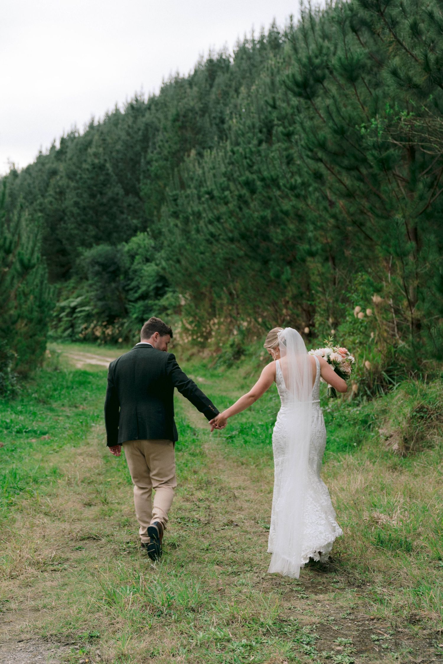 A couple walks hand in hand along a forest trail with evergreen trees in the background.