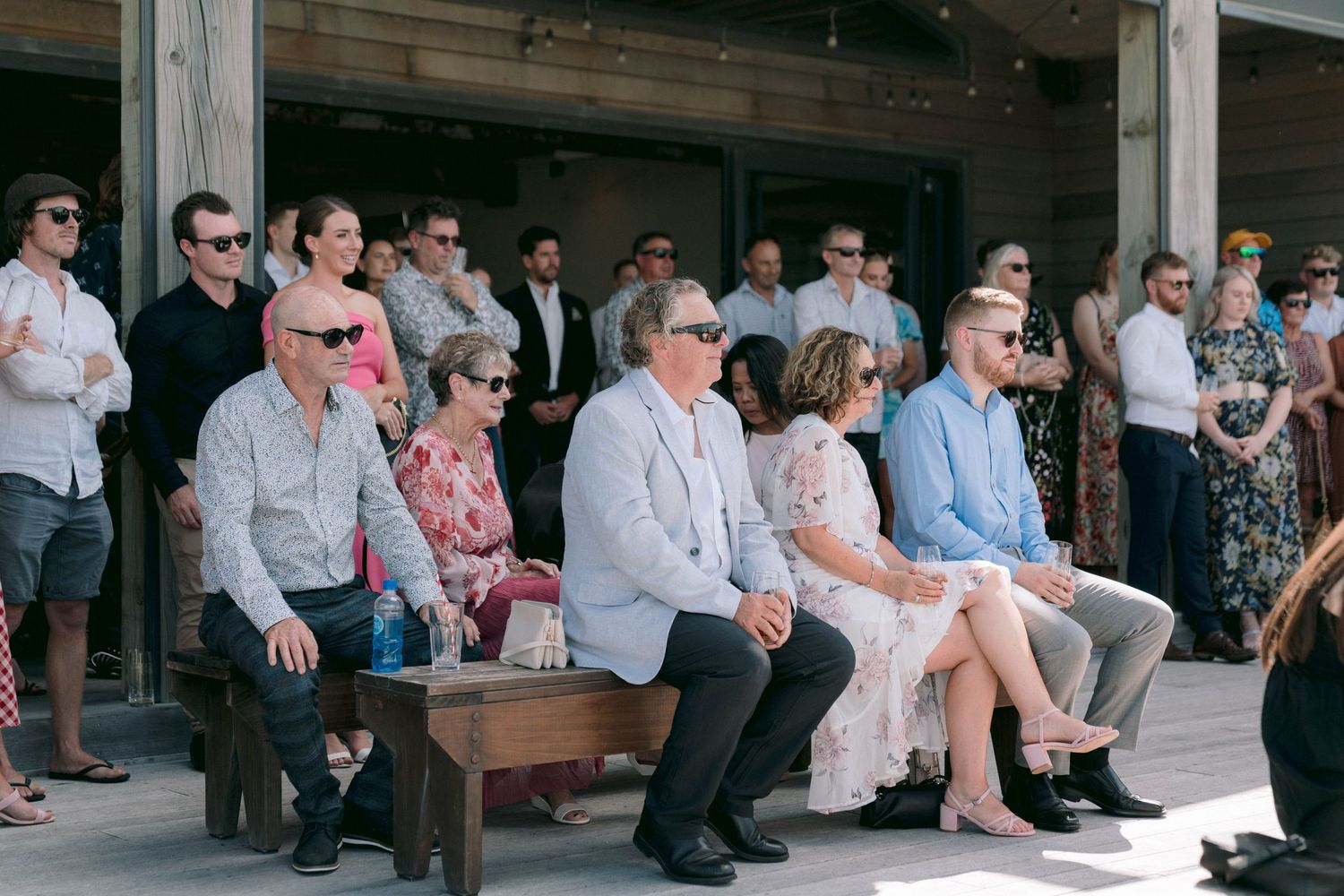 Wedding guests seated outdoors at a rustic venue wearing sunglasses and casual attire while watching ceremony.