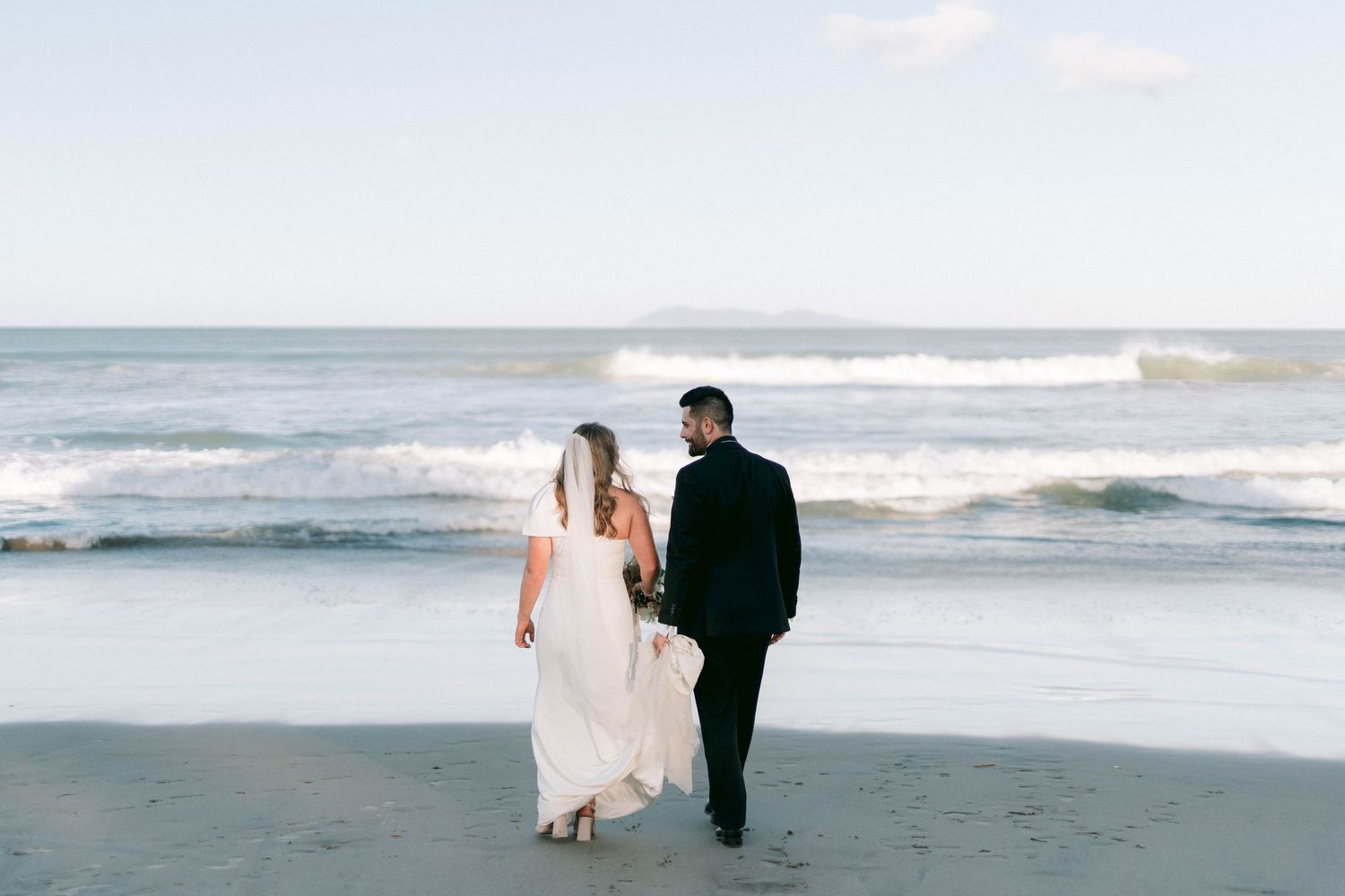 A couple walks together on a beach with waves crashing behind them on their wedding day.