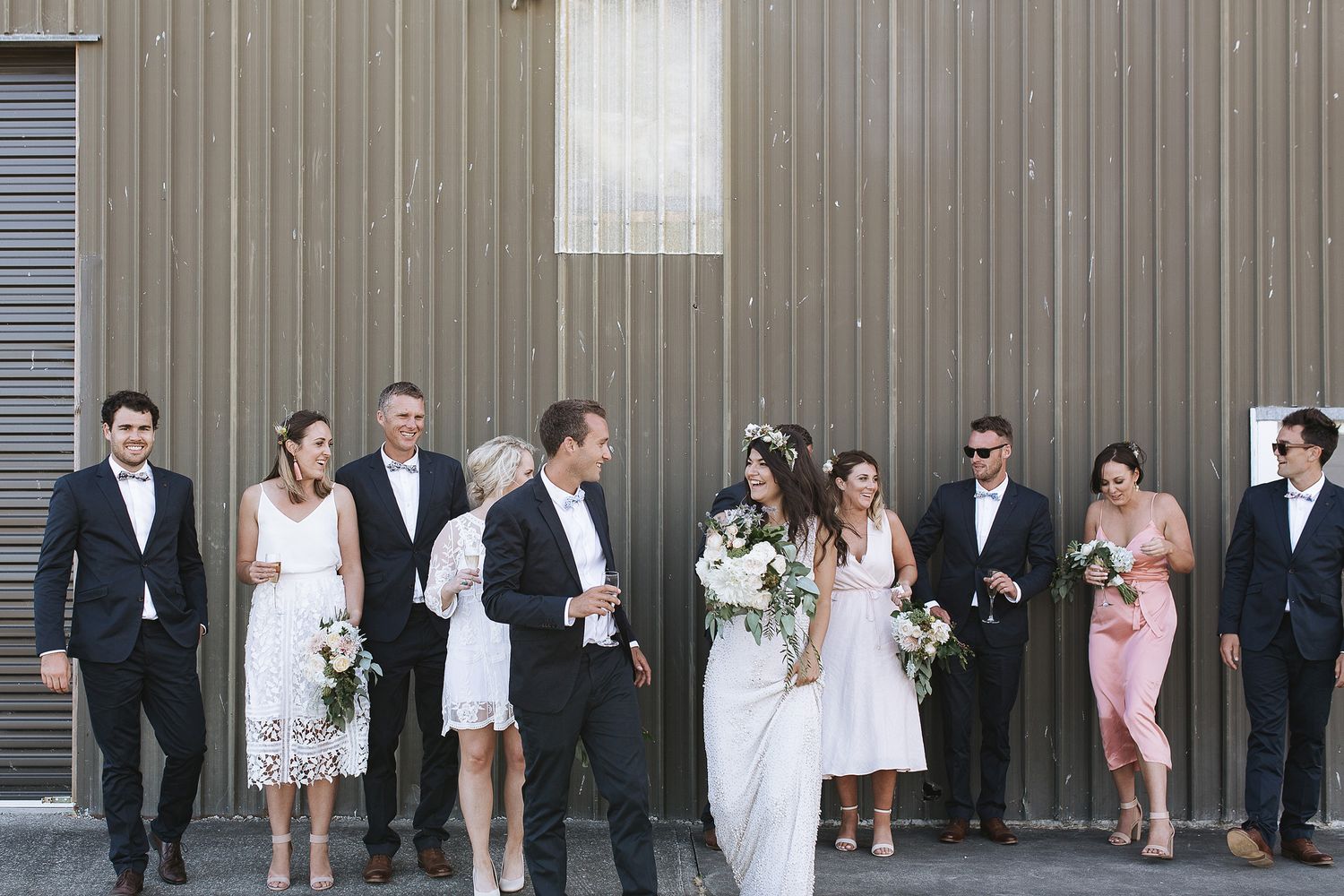 Wedding party stands in line along gray wall for group photo with modern minimal outdoor composition.