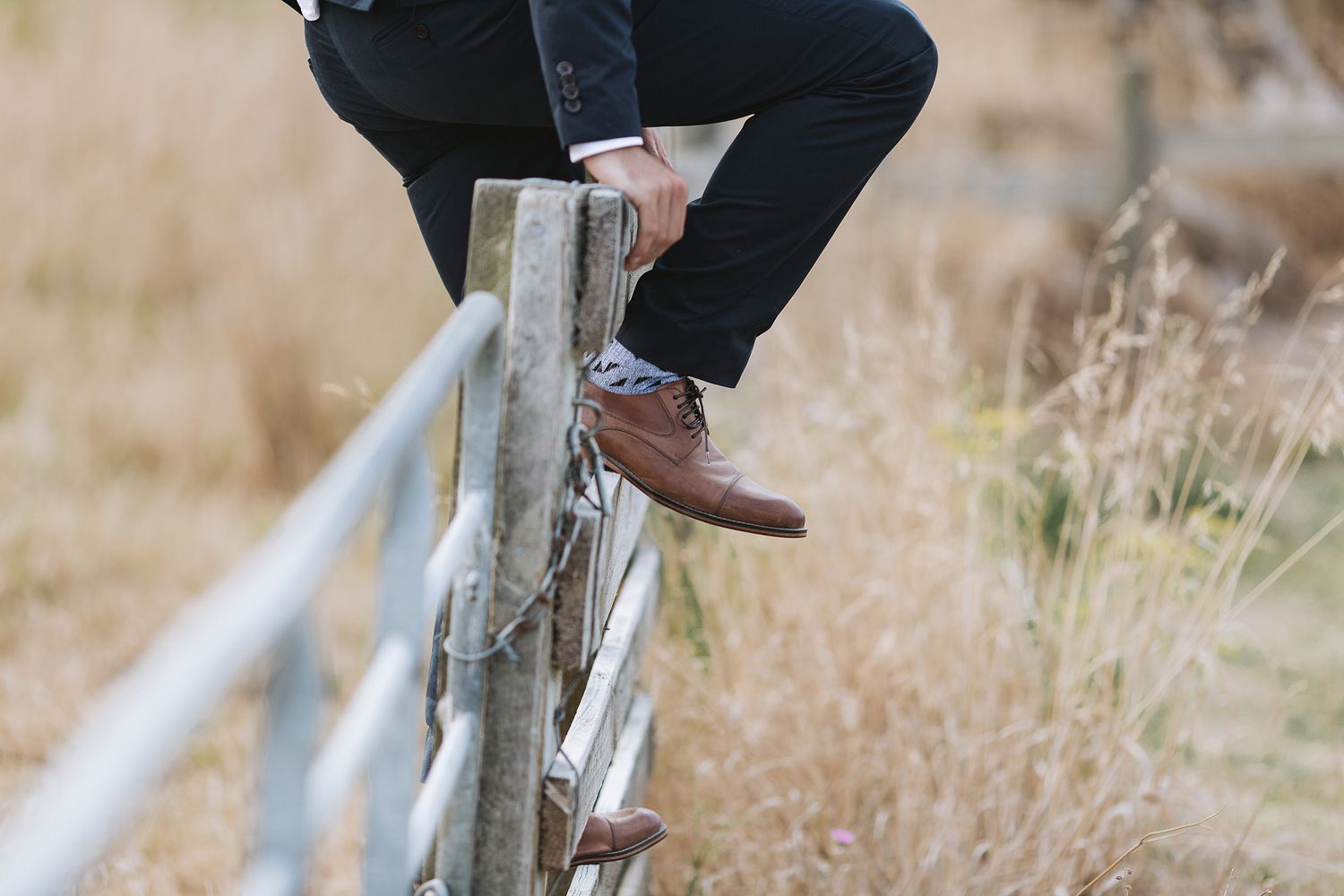 Person wearing brown leather shoes and dark pants sits on a rustic wooden fence surrounded by tall grass.
