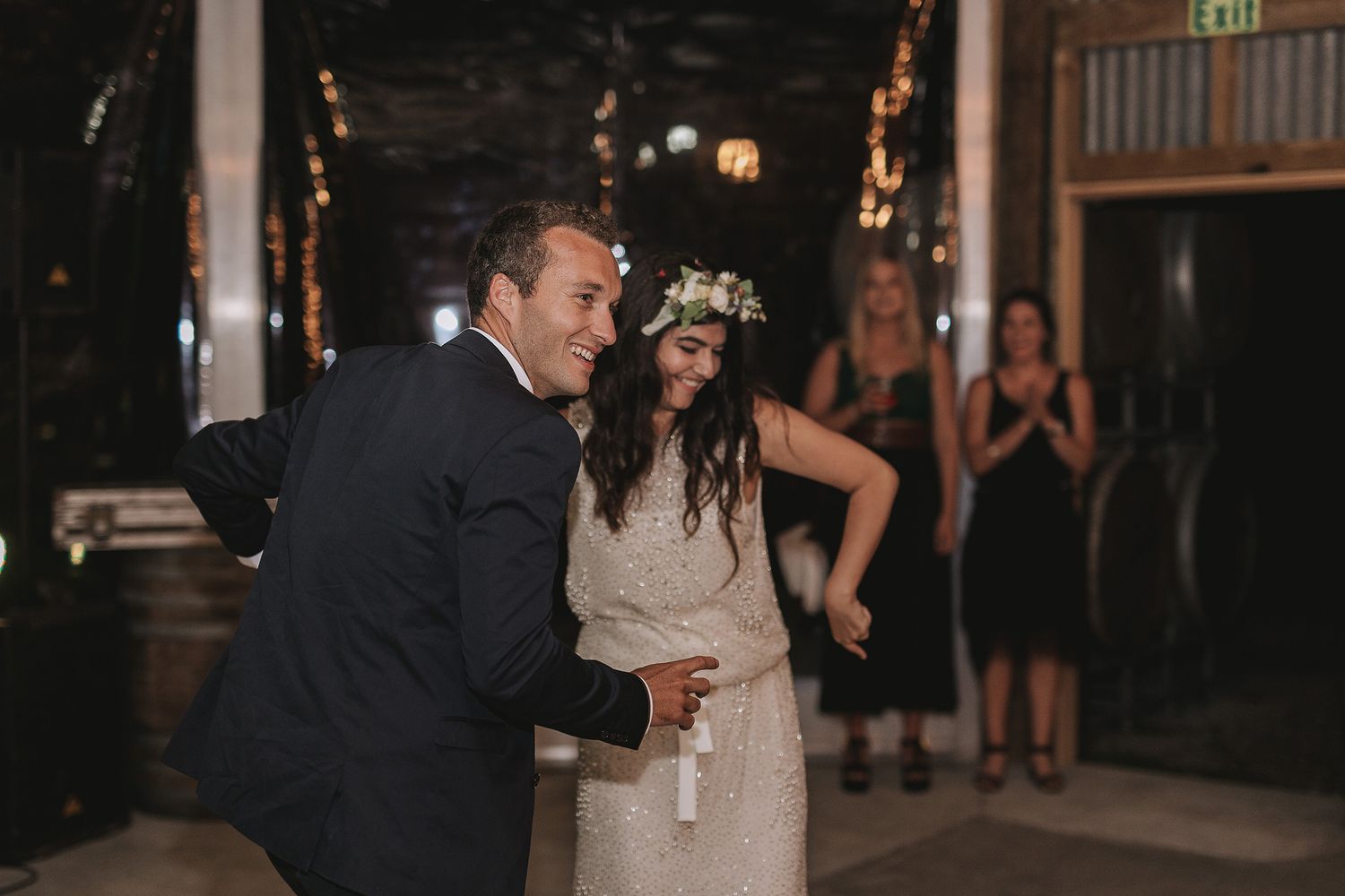 A couple embraces during their first dance at a dimly lit wedding reception celebration with guests watching in the background.