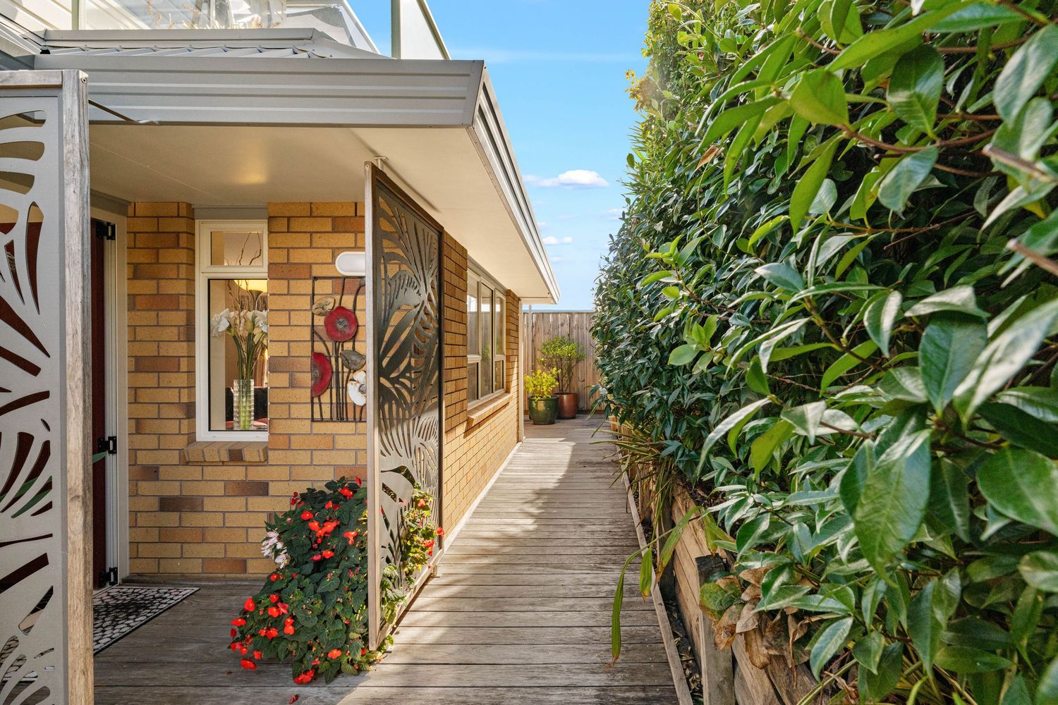 A wide verandah of a brick house with potted red flowers and lush greenery along the walkway.