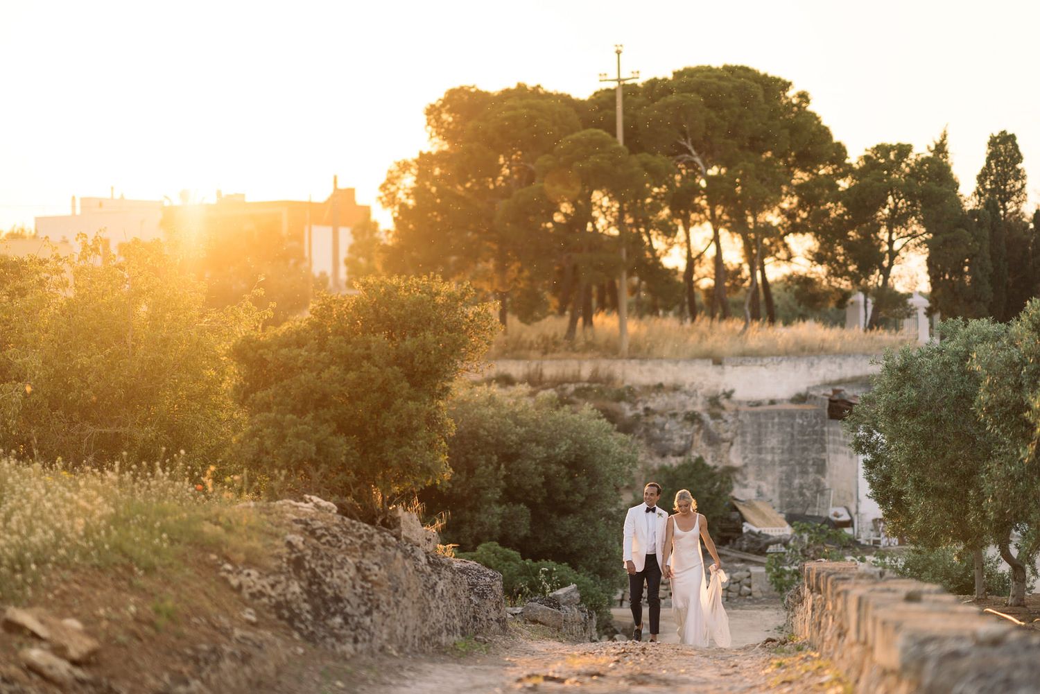 Scenic golden hour wedding photo on stone path with natural Mediterranean landscape and ancient ruins in background.