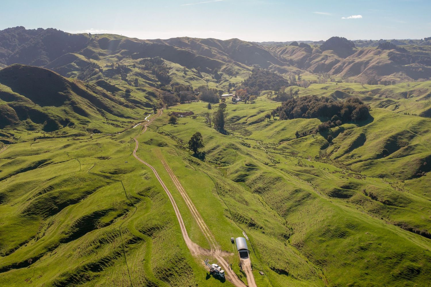 Rolling green hills stretch across a vast landscape dotted with rural farms and winding dirt roads under a blue sky.