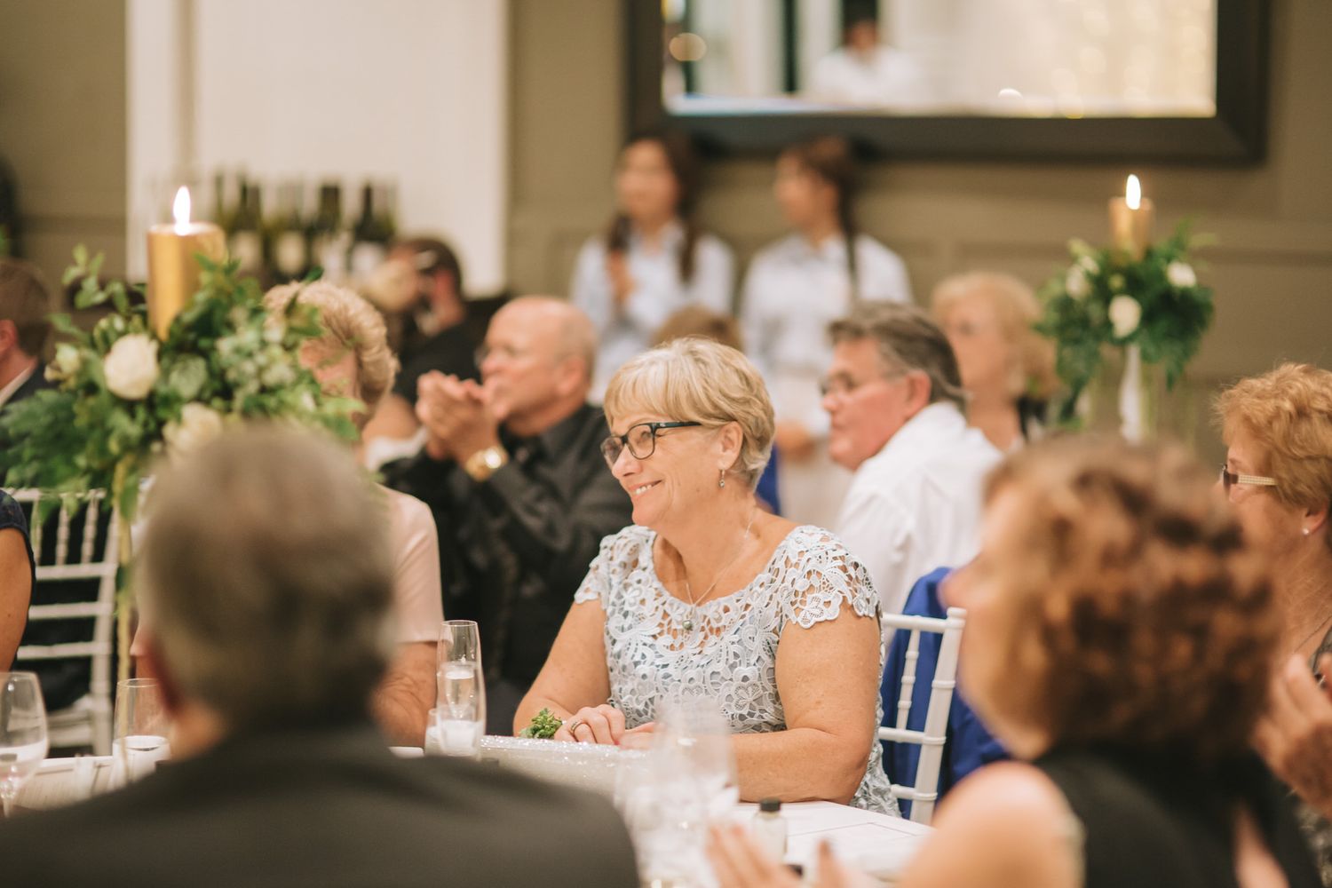 Candlelit wedding reception table with white floral centerpieces and guests seated in elegant chairs at an indoor venue.
