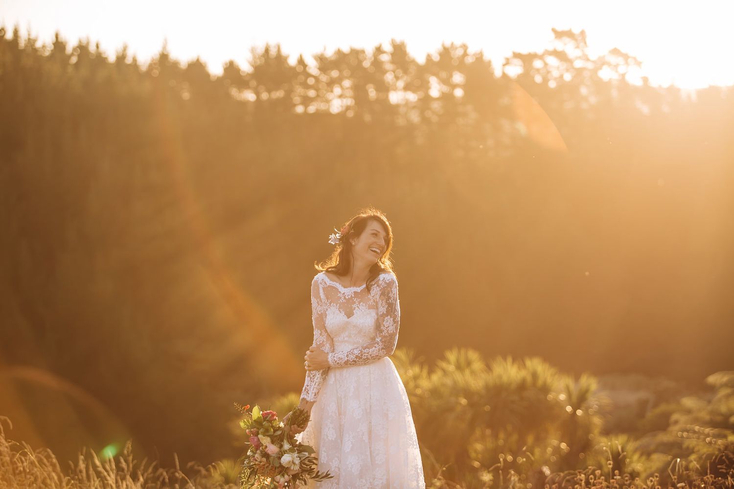 Figure in a flowing white dress stands in a golden-lit meadow at sunset with forest silhouettes in background.