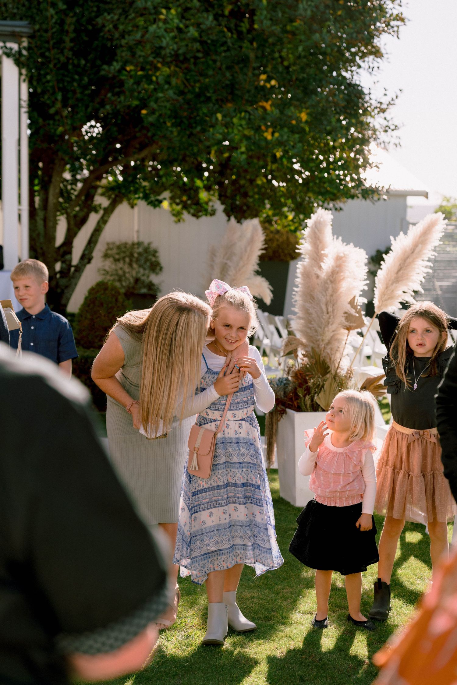 Children playing outdoors in summer attire during a sunny backyard gathering with decorative pampas grass visible.