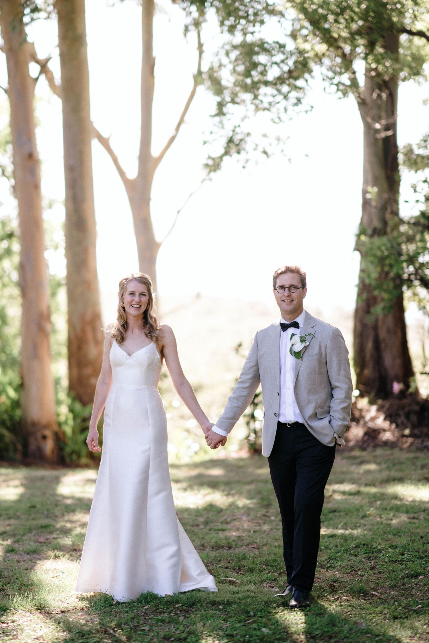 A romantic wedding portrait of a couple holding hands among tall trees in golden sunlight.