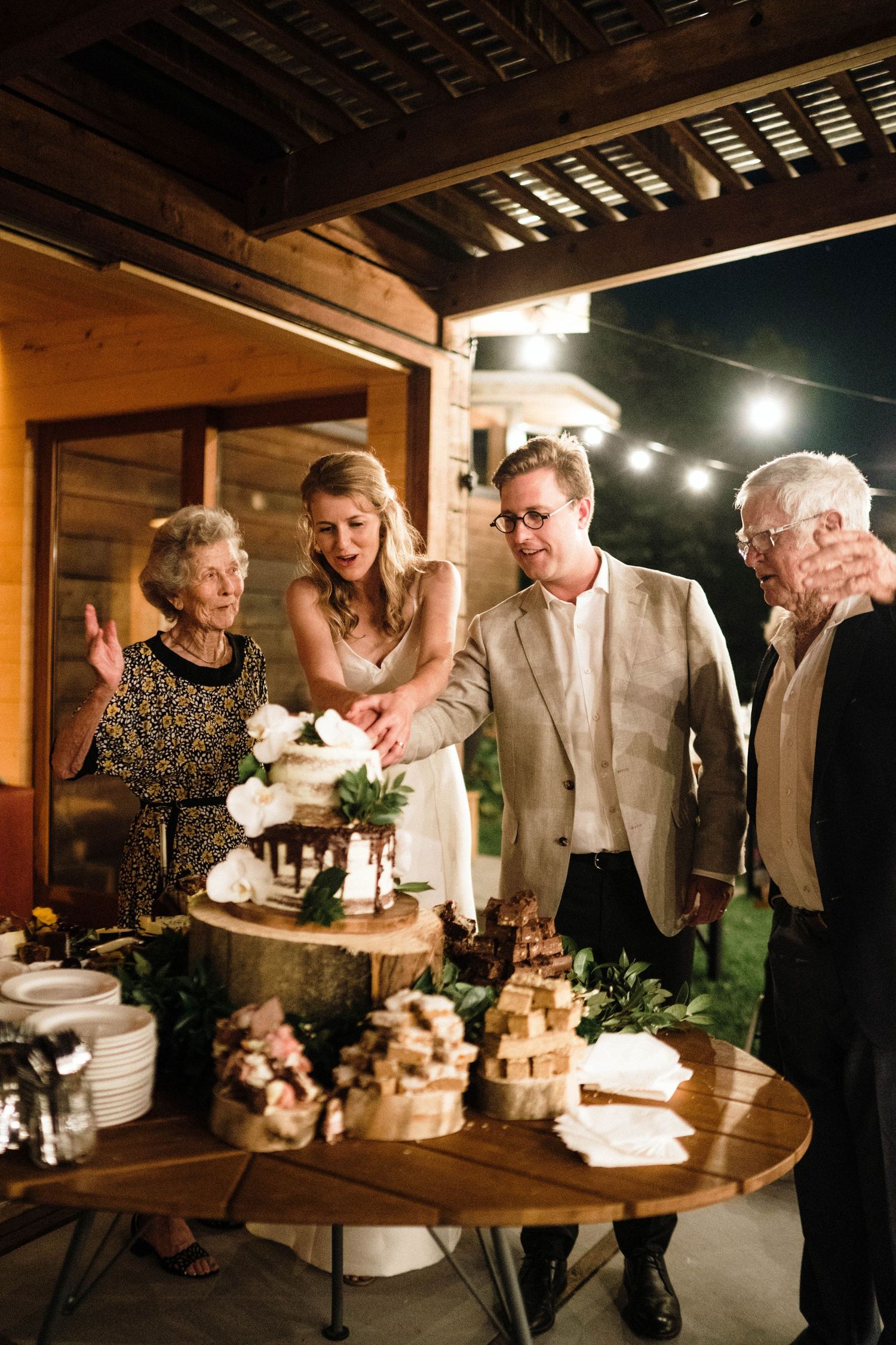 Wedding guests gather around a rustic dessert display with cakes and treats on wooden stands under string lights.
