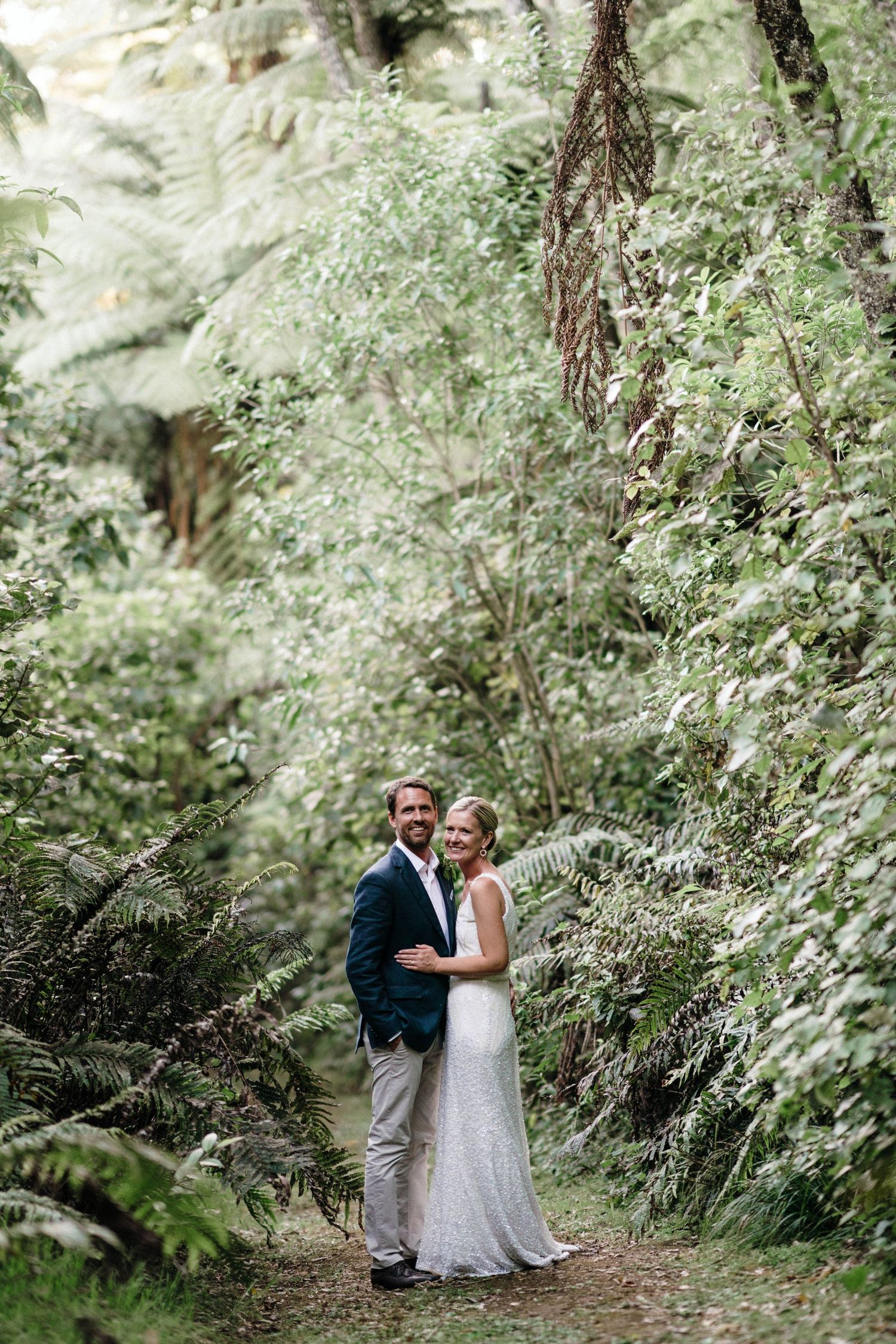 A wedding couple stands on a forest path surrounded by ferns and dappled sunlight.