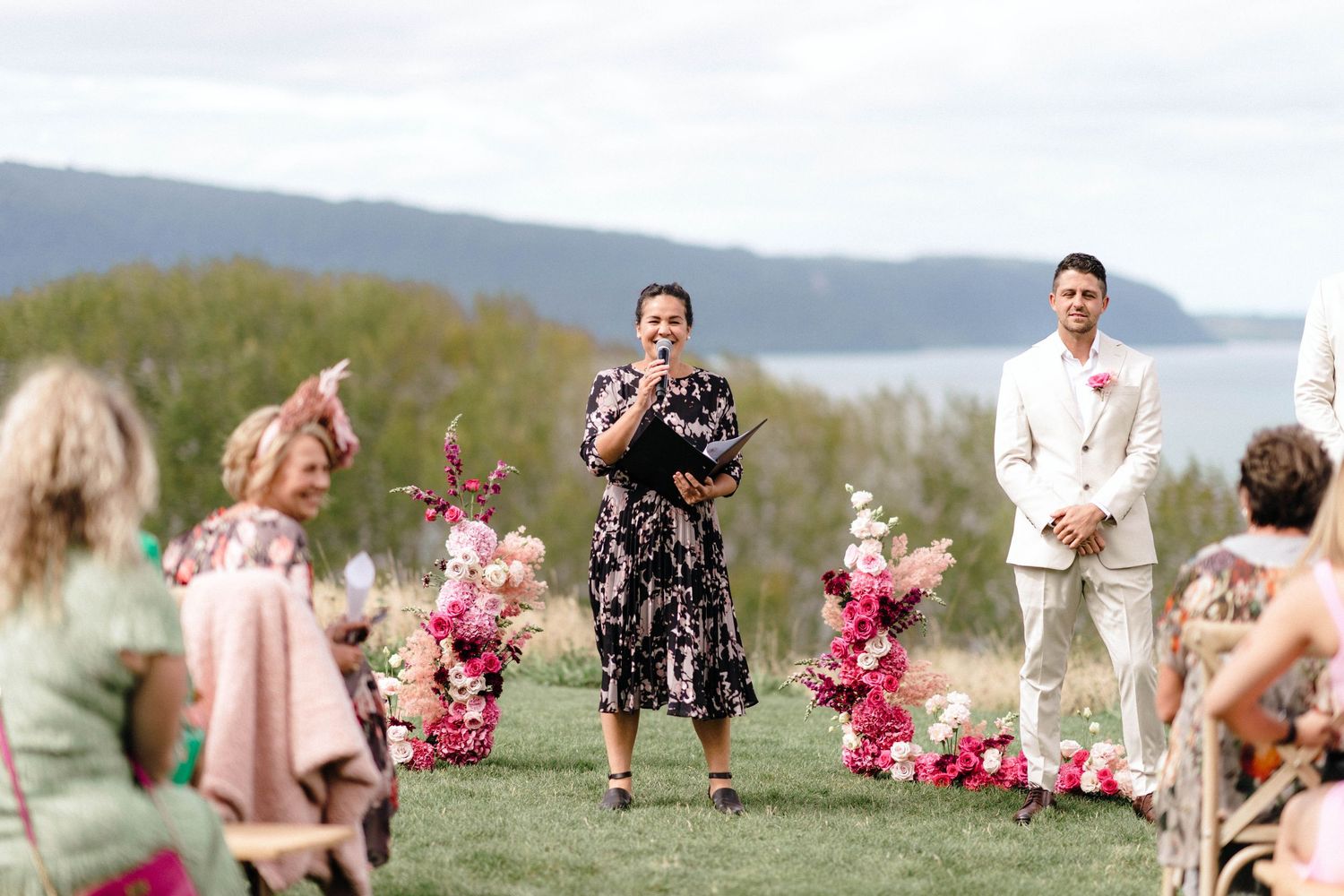 Outdoor wedding celebration on a grassy hilltop with pink floral decorations and scenic mountain views.